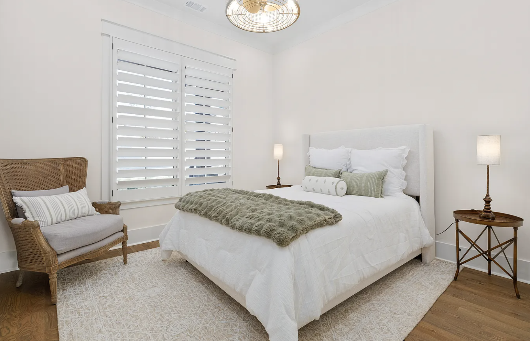 A minimalistic bedroom with a white bed, green accent pillows, and a brown furry throw. There is a wicker chair with a striped pillow, two side tables with lamps, a window with white plantation shutters, and a gold ceiling light fixture. The room has