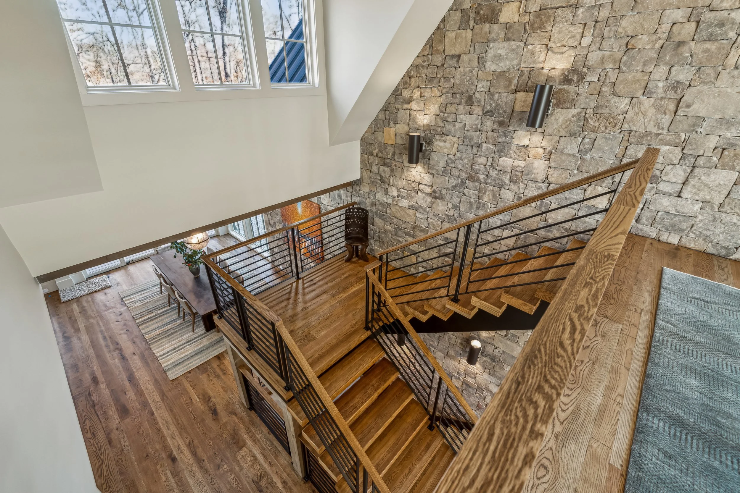 Living room with large windows, stone accent wall, staircase with wooden steps and metal railing, dining table with chairs, area rug, and potted plant.