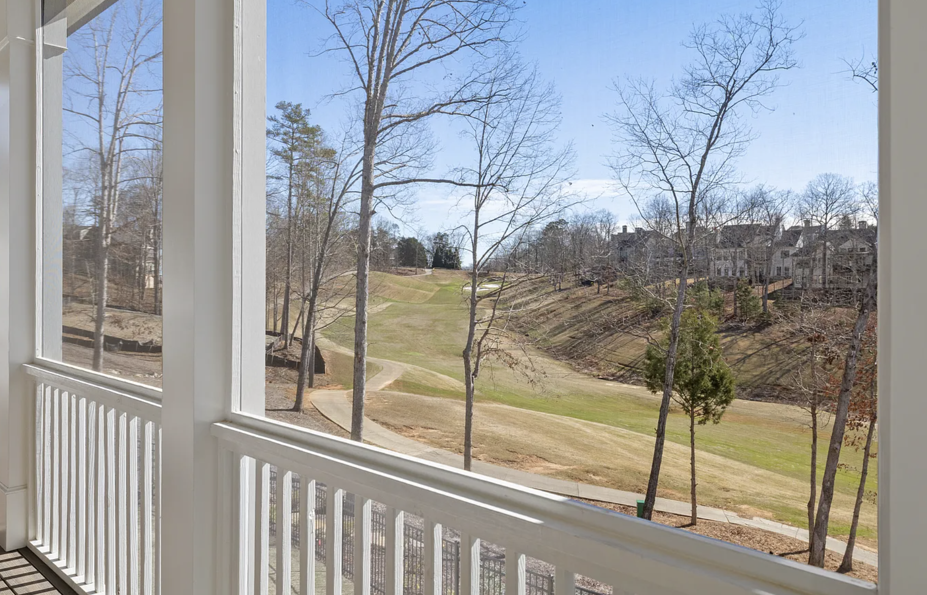View from a porch showing a golf course with a small hill, trees, a pathway, and residential buildings in the distance under a clear blue sky.