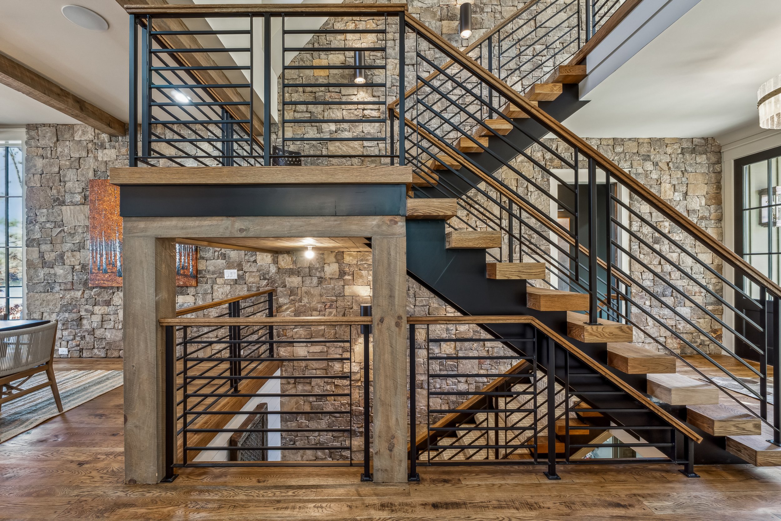 Interior view of a modern staircase with wooden steps and black metal railings, set against a stone accent wall with windows on the right and a wooden floor.