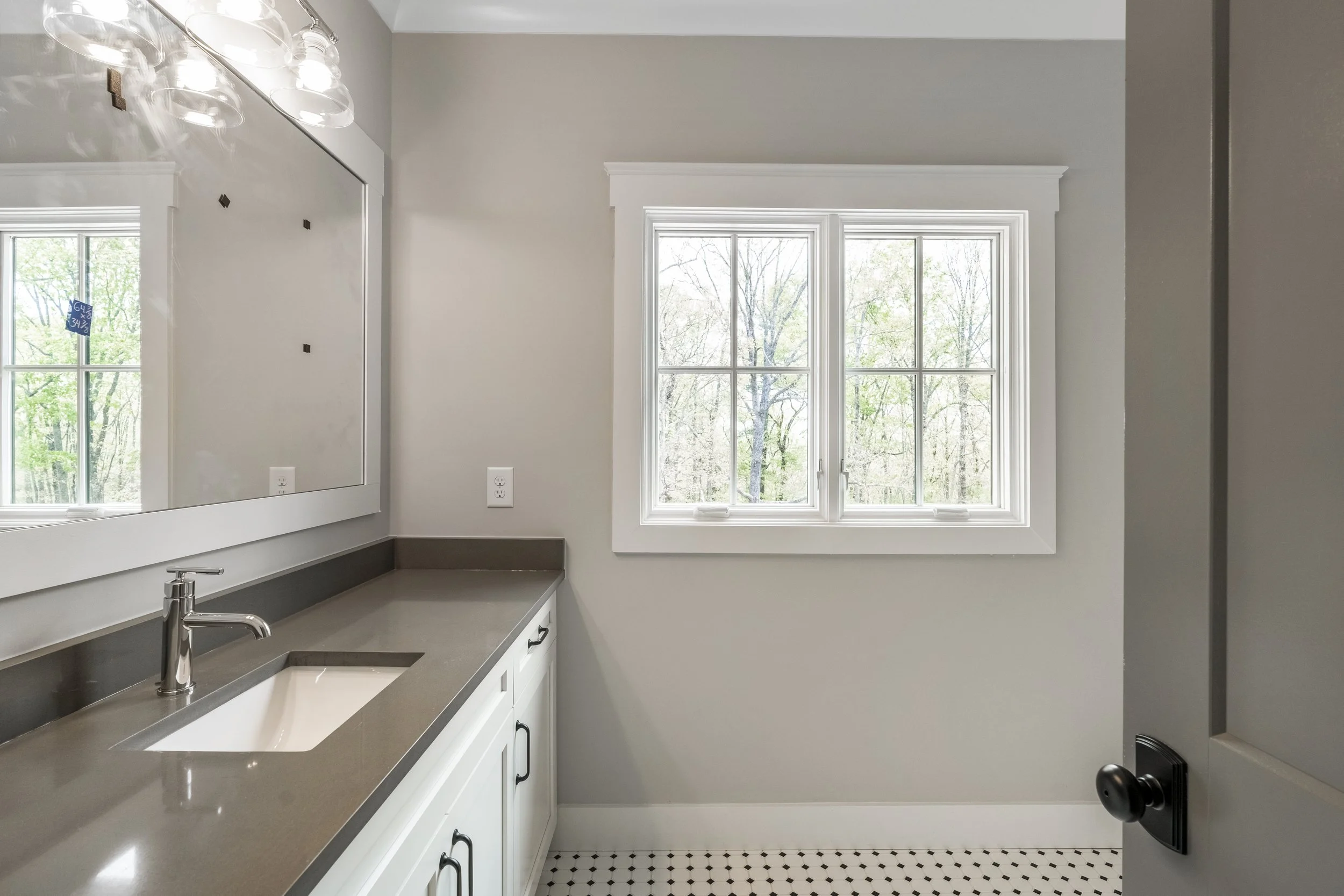 A kitchen with a gray countertop, white cabinets, a stainless steel sink with a modern faucet, and a large window showing trees outside. There is a mirror on the wall reflecting part of the room and bright light fixtures above the mirror.
