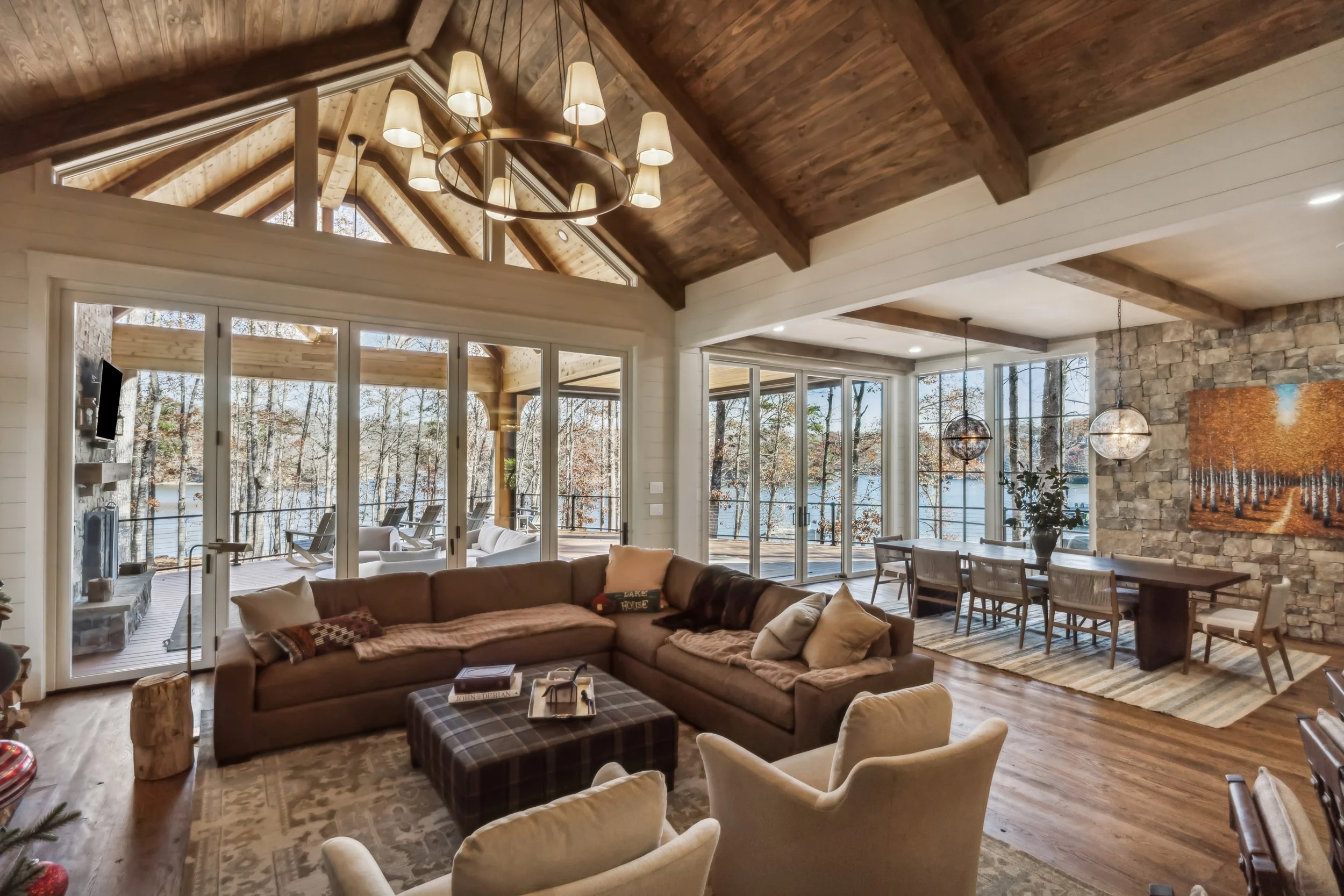 A spacious living room with large windows facing a wooded landscape. It features a vaulted wooden ceiling, a stone accent wall, a sectional sofa, armchairs, a plaid ottoman, and hanging spherical pendant lights.