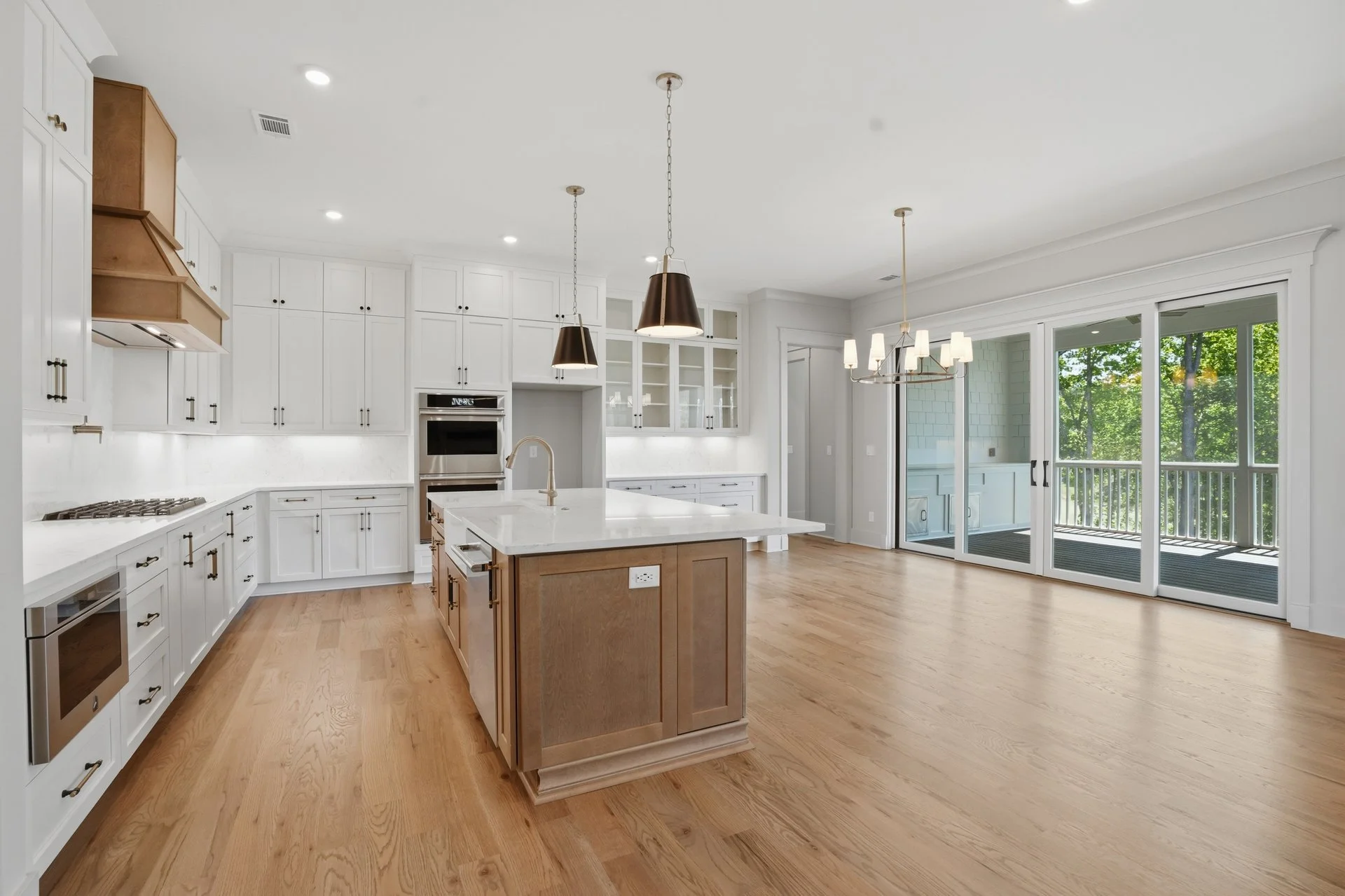 Modern kitchen with white cabinets, a central island with a wooden base, stainless steel appliances, hardwood floors, and large sliding glass doors leading to an outdoor balcony with greenery.