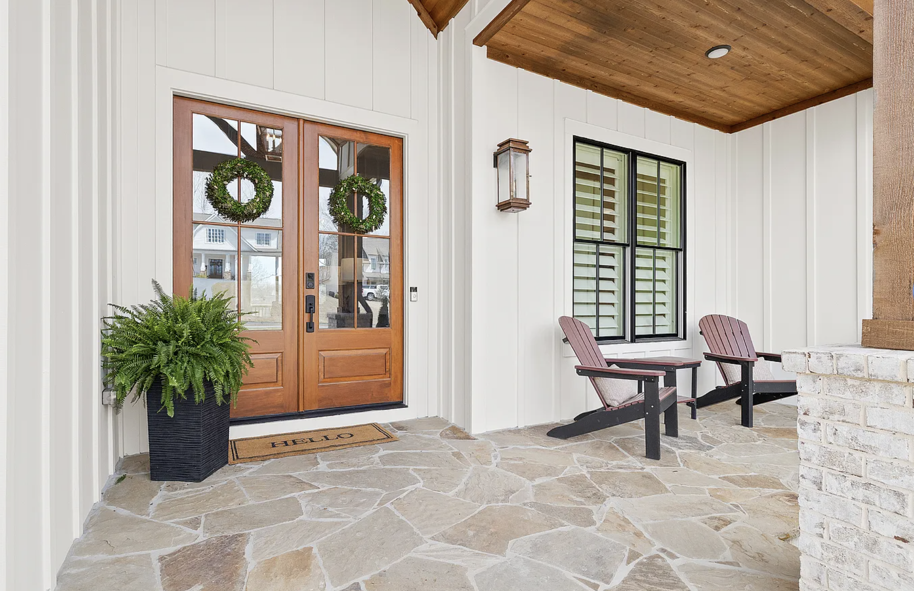 Front porch area with a wooden door decorated with two green wreaths. A large potted fern plant is on the left, and two red chairs with black frames are on the right. The floor is made of irregular stone tiles, and there is a brick half-wall on the r