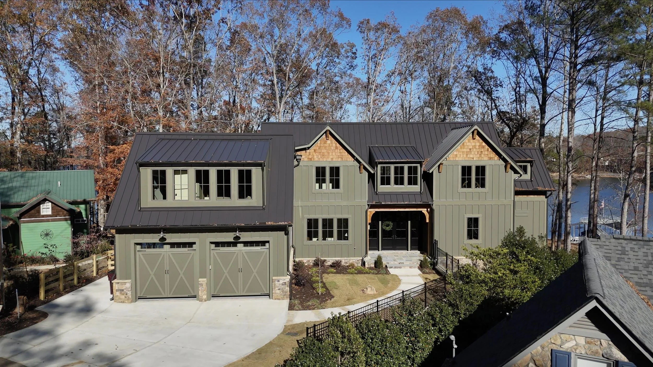 A modern two-story house with gray vertical siding, large windows, and a dark metal roof, situated in a wooded area with autumn trees and a water body visible in the background.