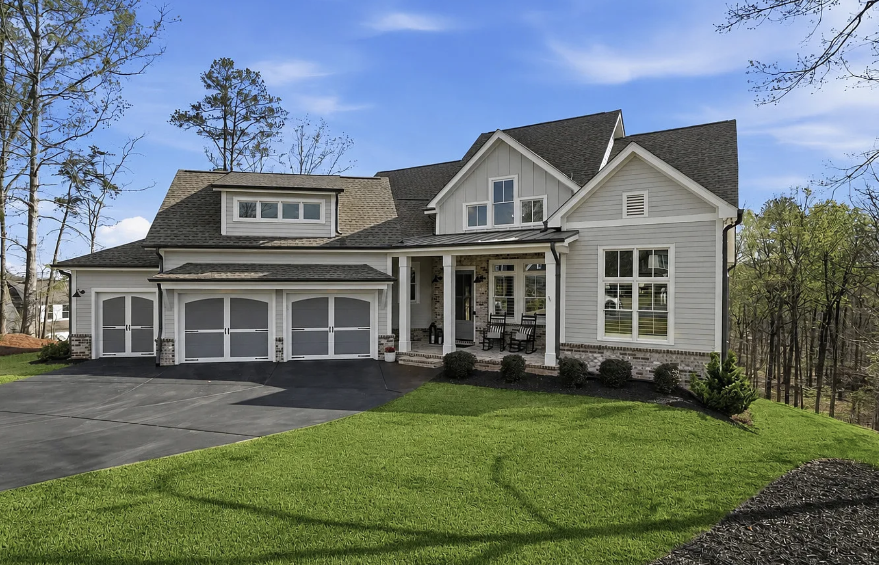 A modern two-story house with white siding, a three-car garage, a front porch with chairs, and a well-maintained lawn, set against a backdrop of trees and a blue sky.