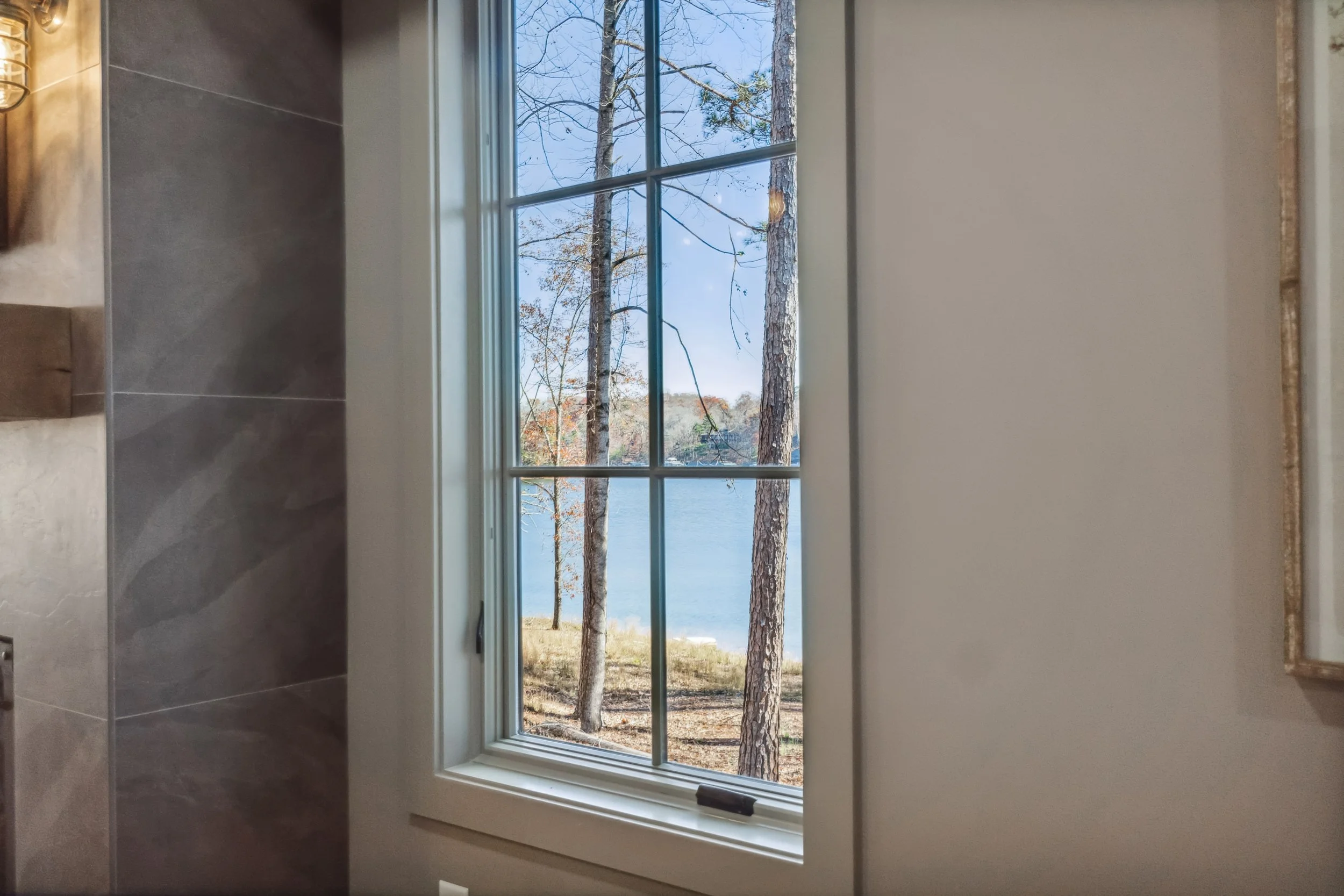 A view through a window showing trees and a body of water outside, with the photo taken from inside a room that has beige walls and gray tile flooring.