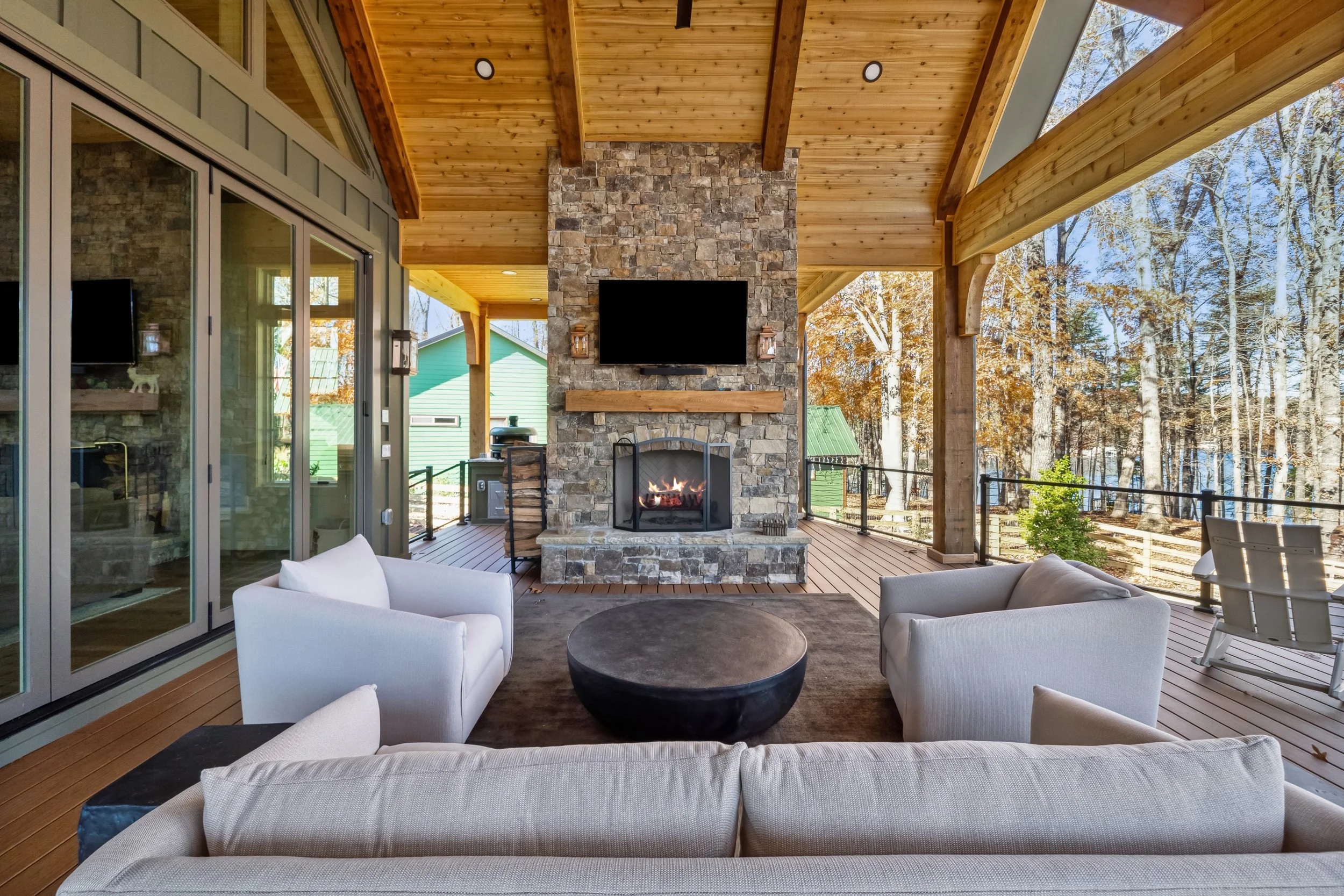 Outdoor living area with a stone fireplace, mounted flat-screen TV, and a wooden ceiling, surrounded by trees and a deck.