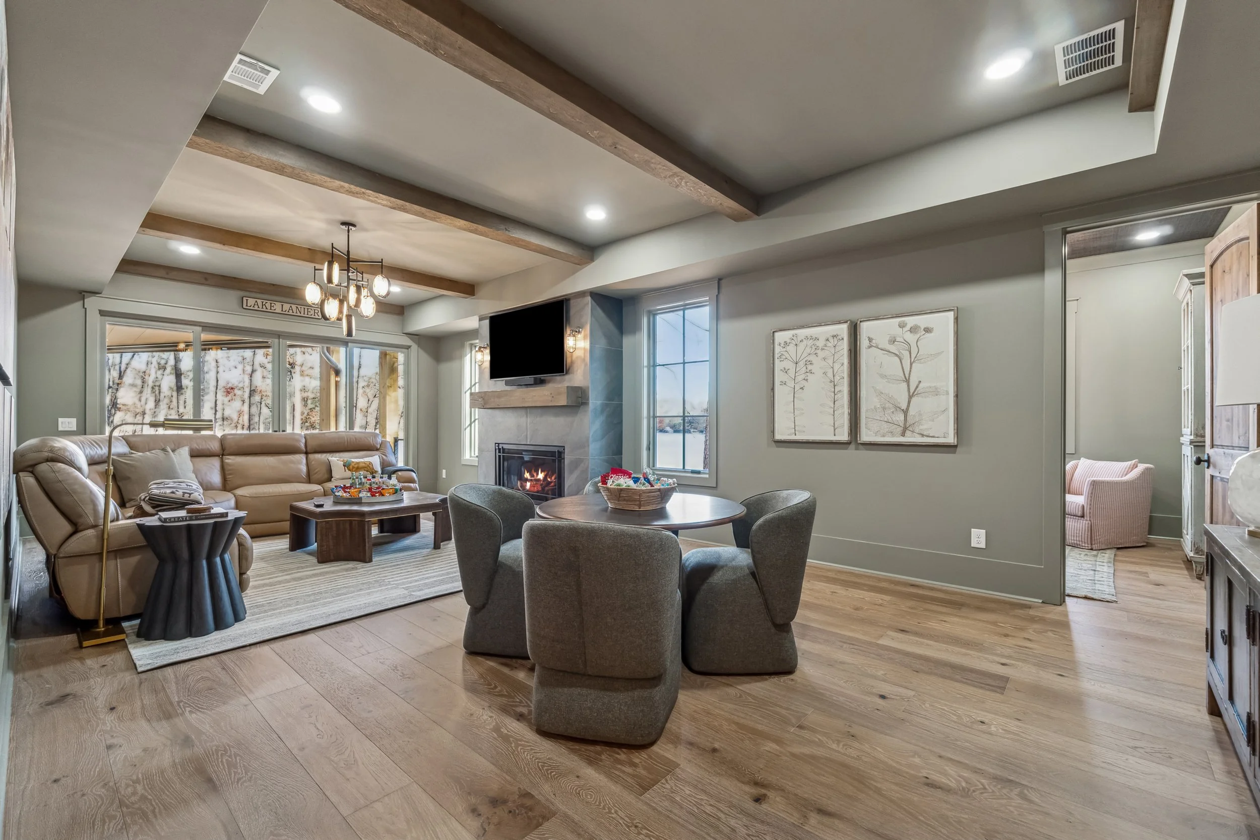 Living room with beige leather sofa, armchair, round wooden table with chairs, fireplace, TV on wall, wood beams on ceiling, large windows, contemporary decor.