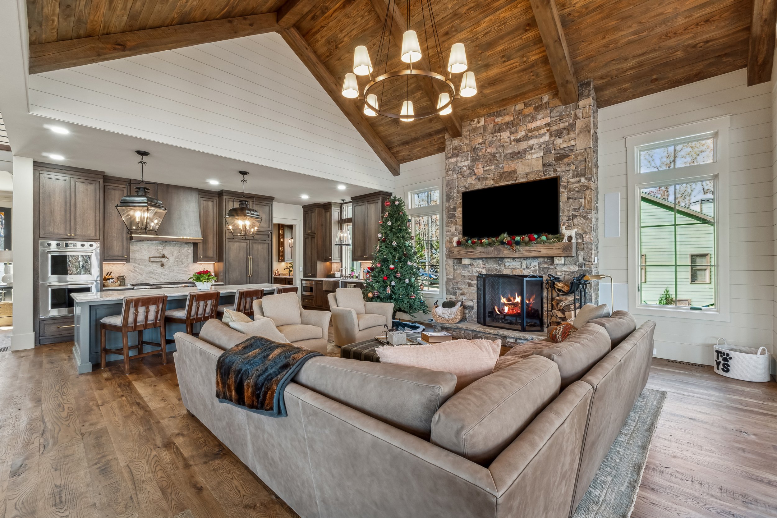 Living room with a high wooden vaulted ceiling, a stone fireplace with a TV above, a Christmas tree, and a beige sectional sofa.
