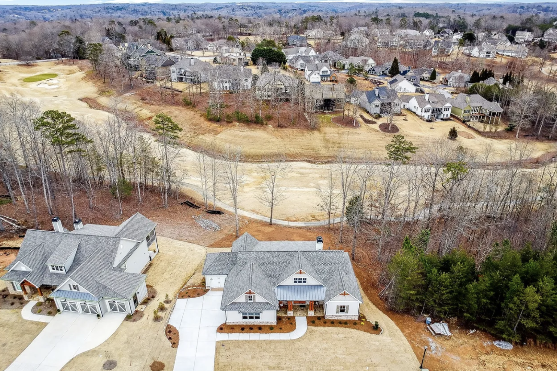 Aerial view of a suburban neighborhood with several new houses, grassy yards, and wooded areas, with an open patch of land or dirt lot in the foreground and more houses in the distance.