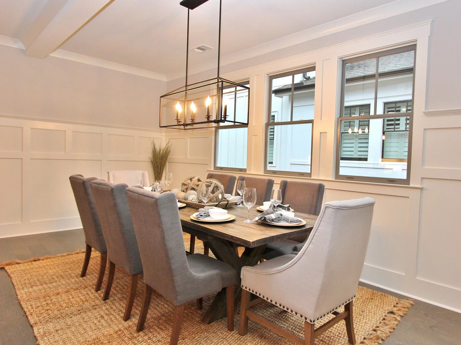 A dining room with a wooden table set with plates, glasses, and napkins, surrounded by various upholstered chairs, a modern light fixture hanging from the ceiling, large windows on one wall, and a woven rug on the floor.