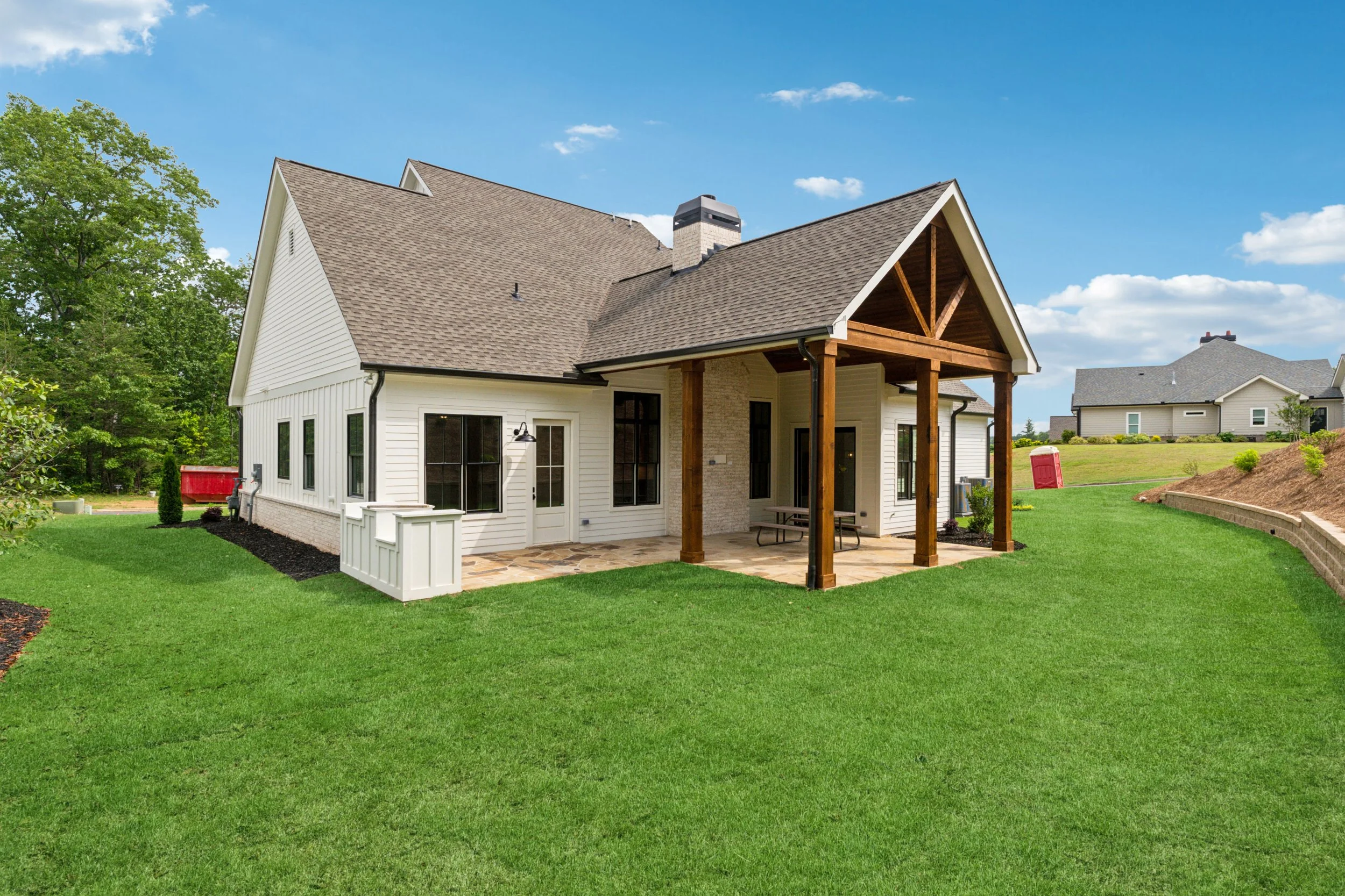 Backyard of a modern house with a covered patio, green grass, and neighboring houses in the background under a blue sky with clouds.