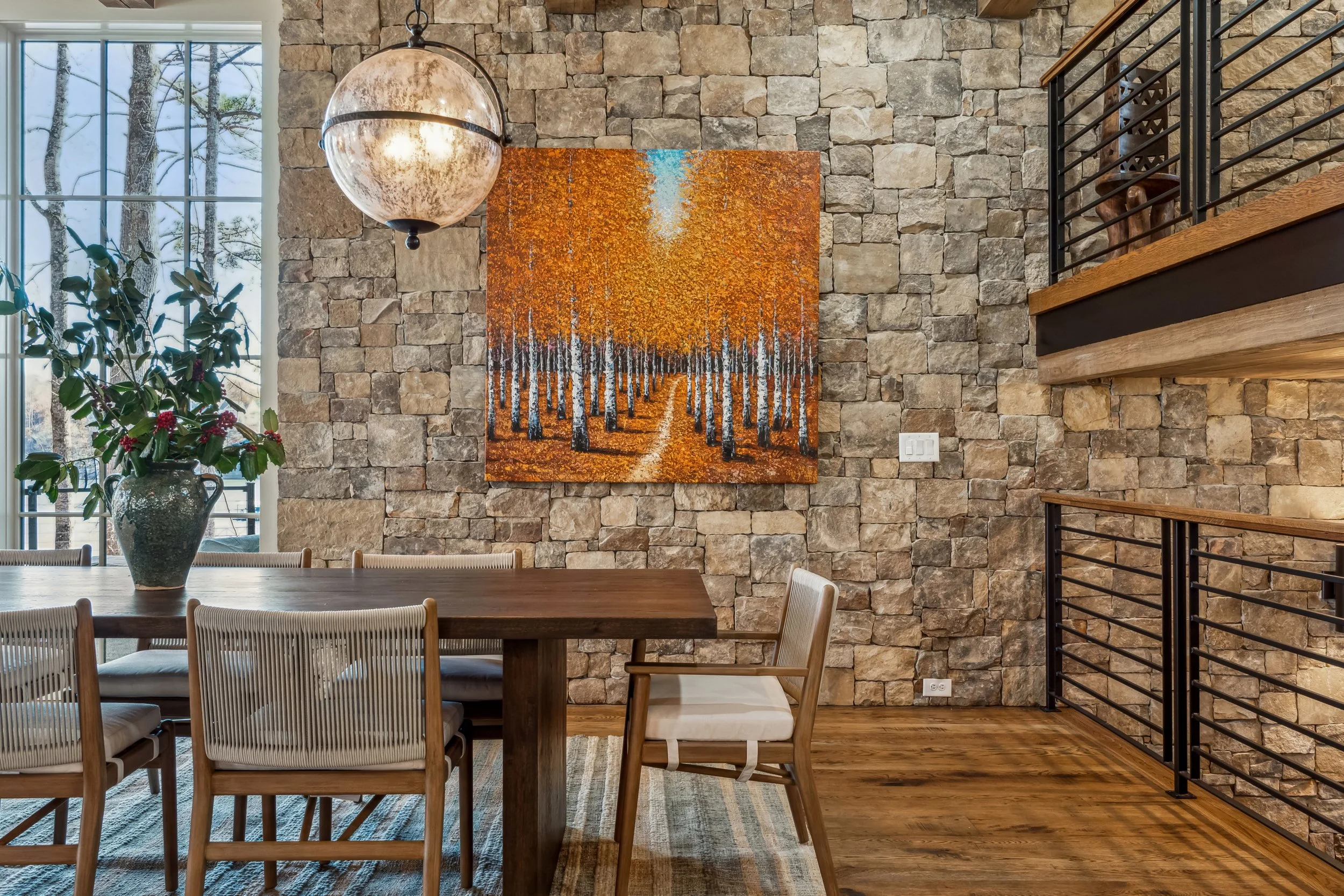 A dining area with a stone accent wall, a large window with a view of trees, a painting of a forest with orange leaves, and a hanging spherical light fixture. There is a table with several white chairs and a vase with green leafy plants and red berri