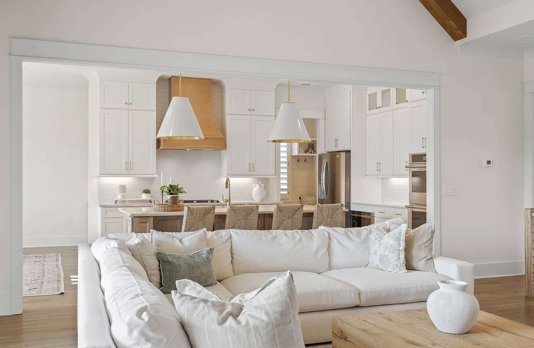 Open-concept living room with white sectional sofa, pillows, and wooden coffee table, overlooking a kitchen with white cabinetry, gold hardware, pendant lights, and a wooden range hood.
