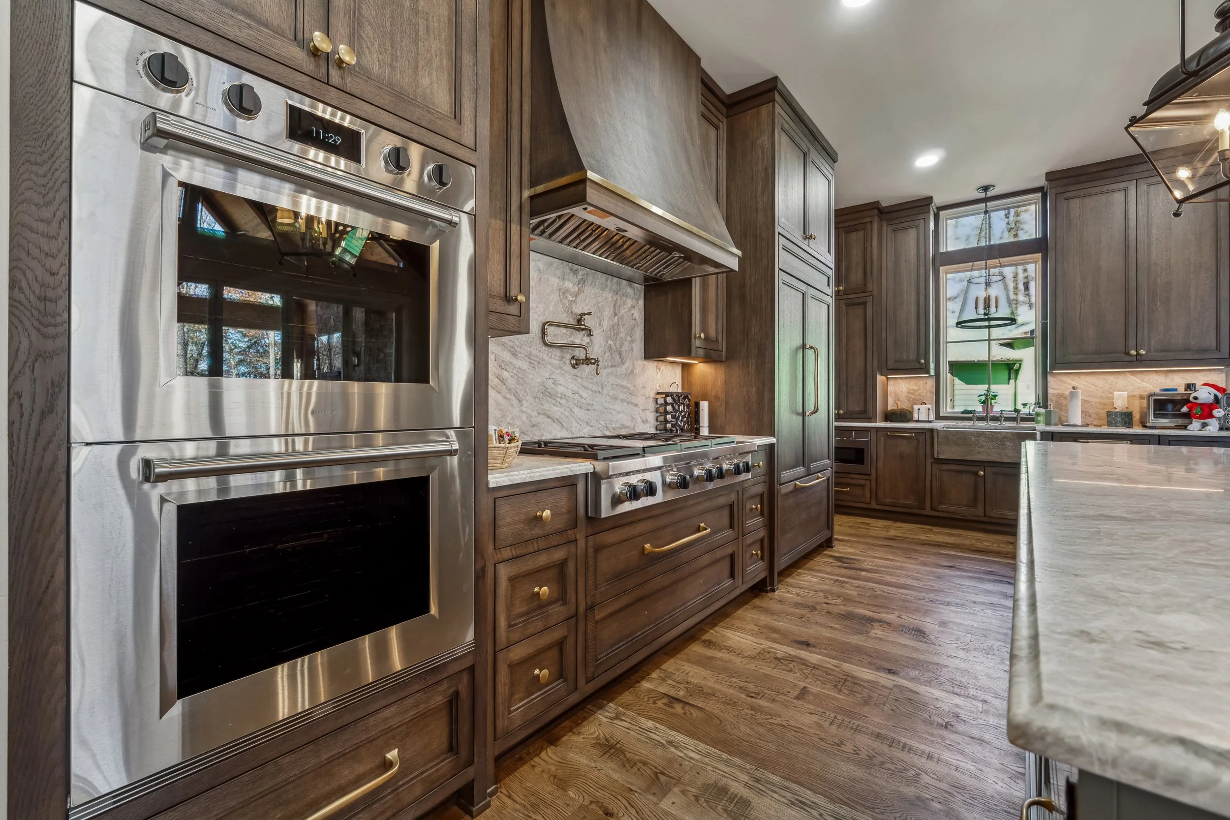 A modern kitchen with dark wooden cabinets, stainless steel appliances including an oven and range, marble countertops, and a window with a view of trees outside. There is a Christmas-themed decoration on the countertop.