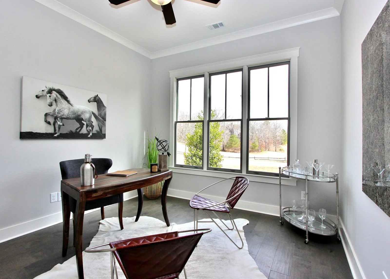 A bright room with a large window showing a view of trees and a fenced yard. The room has a wooden desk with a black chair, a decorative sculpture, a potted plant, and a bottle. Two modern chairs, a white rug, a bar cart with glassware, and a black a