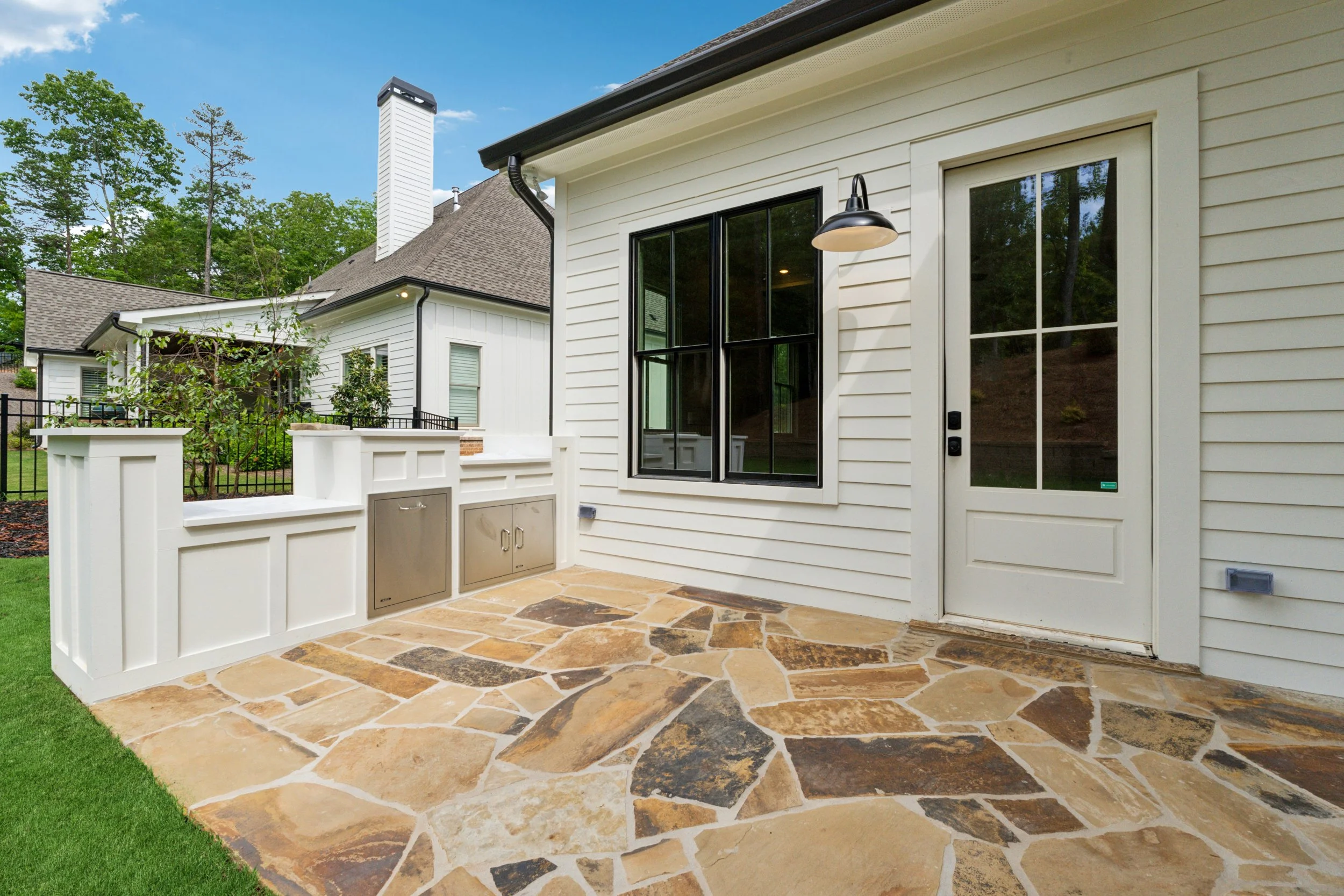 Back porch with stone flooring, white siding, black-framed glass door, and outdoor wall light fixture. In the background are neighboring houses with roofs, trees, and a clear blue sky.