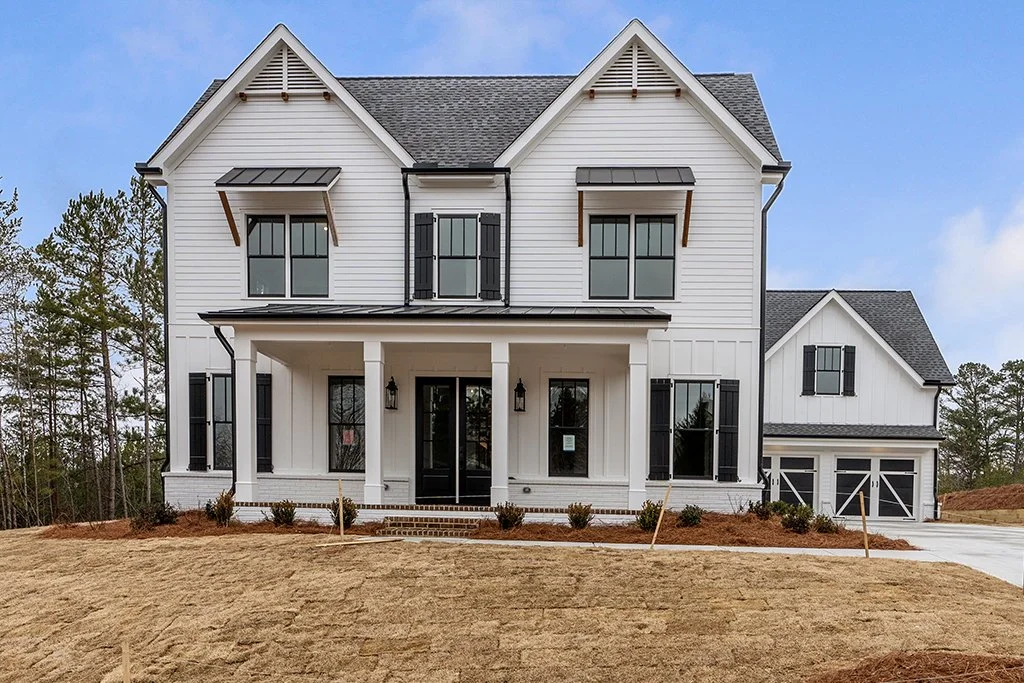 New two-story white house with black shutters, front porch, and attached garage, surrounded by a lawn and trees in the background.