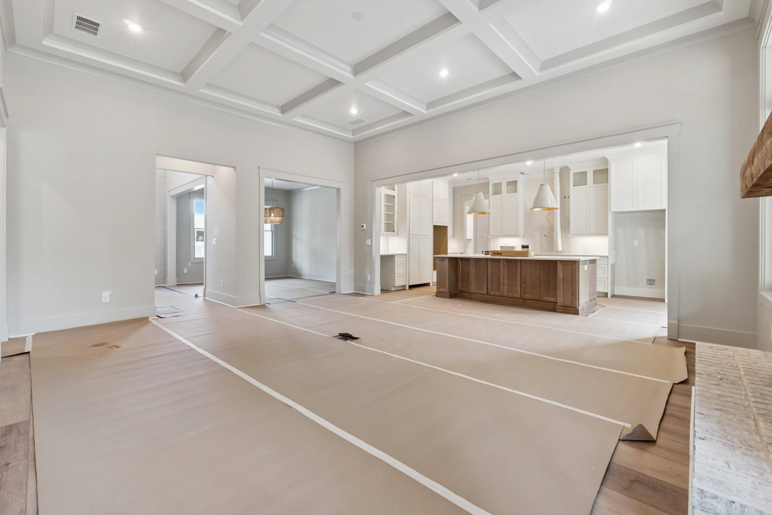 Interior of a house under construction, showing a spacious open-plan kitchen and living area with white walls, a coffered ceiling, and a kitchen island with pendant lights.