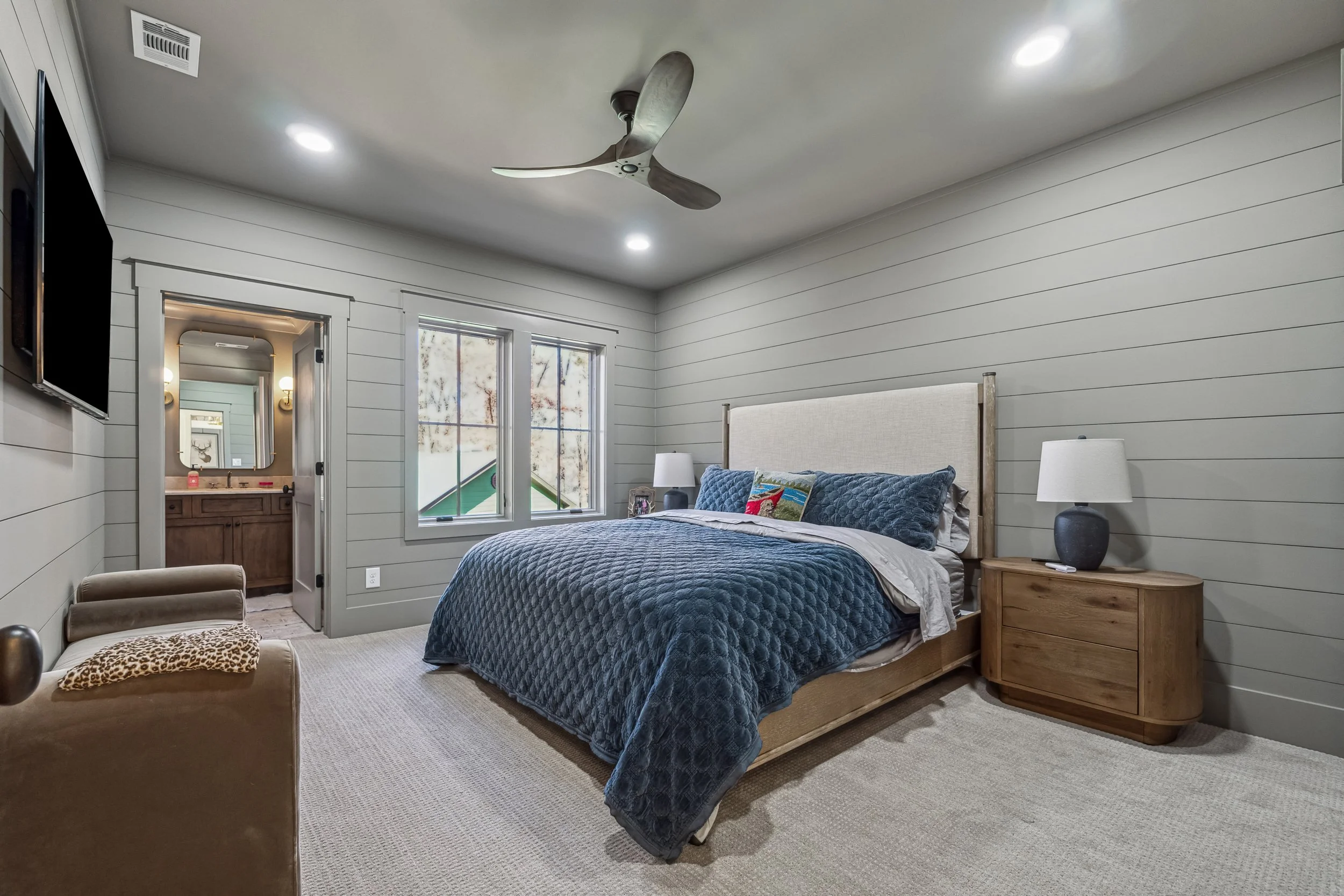 A bedroom with a wooden bed frame, navy quilt, white pillows, and two nightstands with lamps. There are windows and a doorway leading to a bathroom with a mirror and vanity.