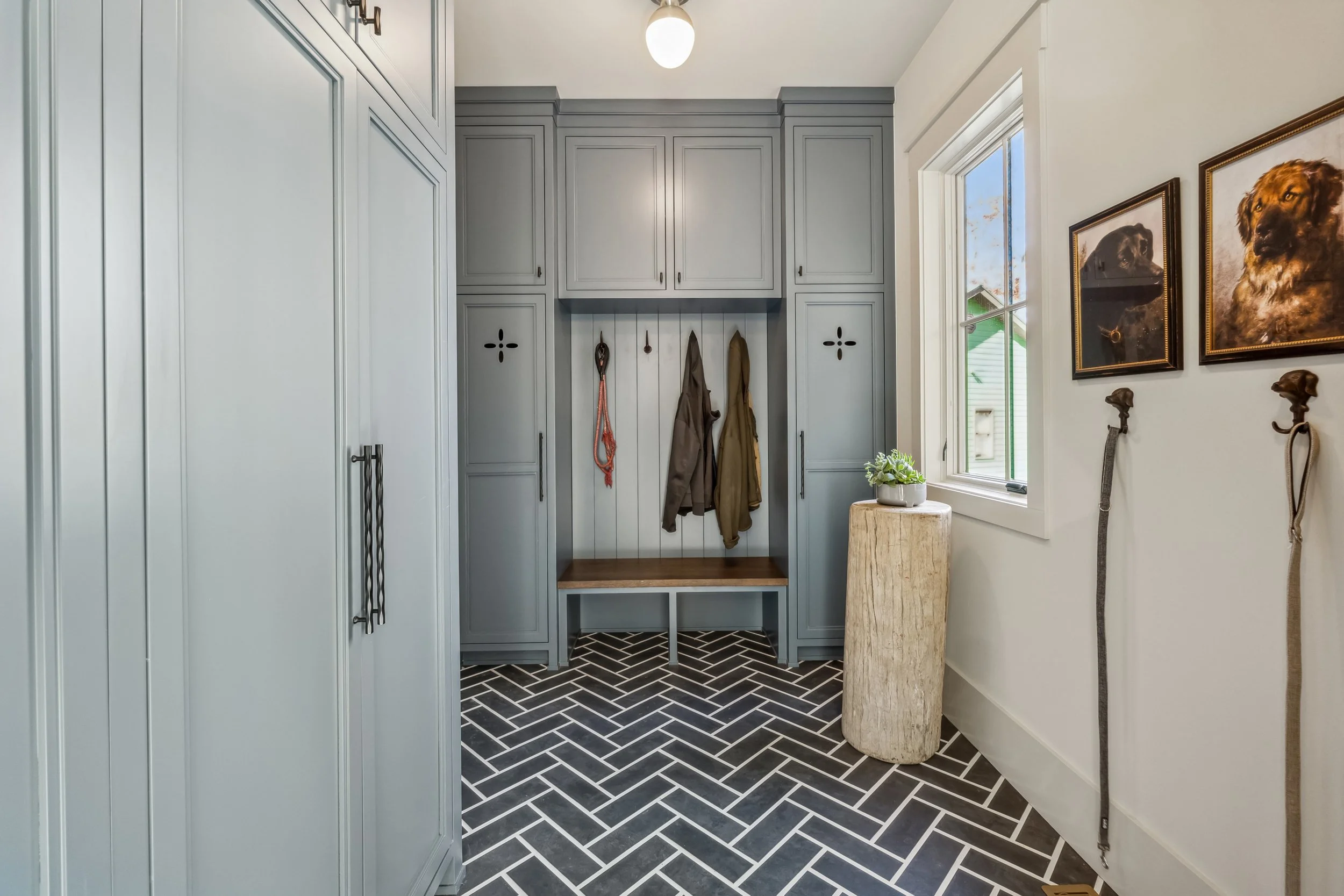 A mudroom with light blue cabinets, black and white herringbone tile floor, a window with outside view, and framed dog portraits on the wall.