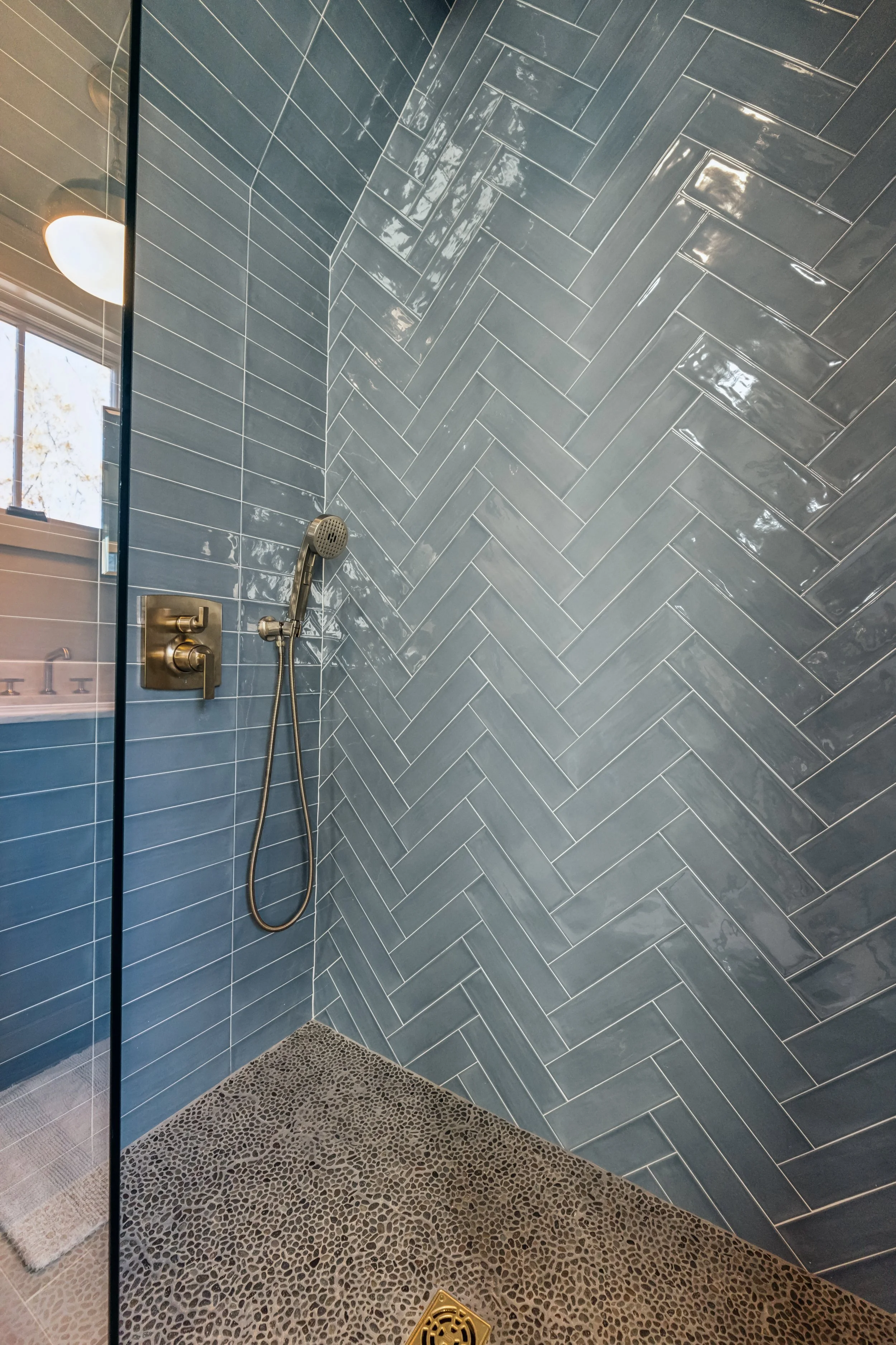 A modern walk-in shower with blue rectangular tiles arranged in a herringbone pattern on the walls, a brushed gold showerhead, and a textured pebble stone floor with a metal drain.