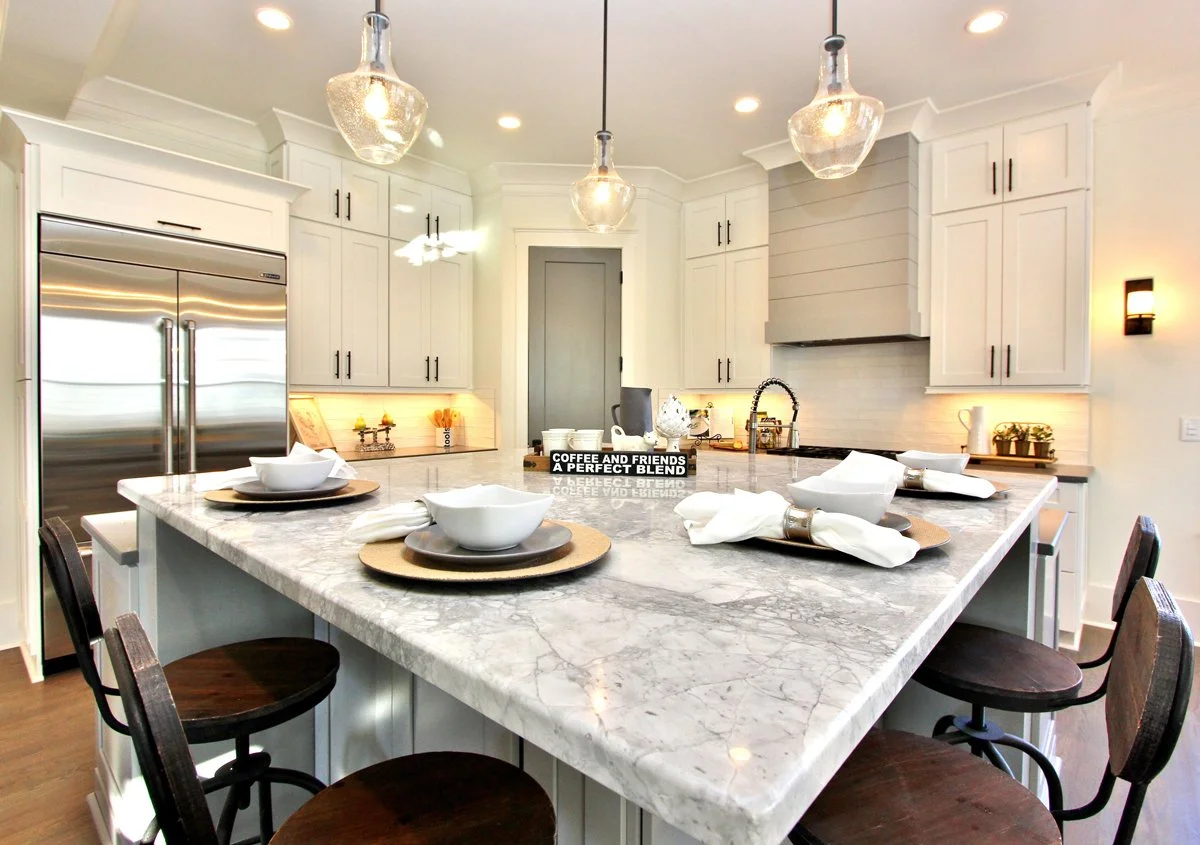 Modern kitchen with white cabinets, a large marble island with place settings, pendant lights, stainless steel refrigerator, and decorative accents.