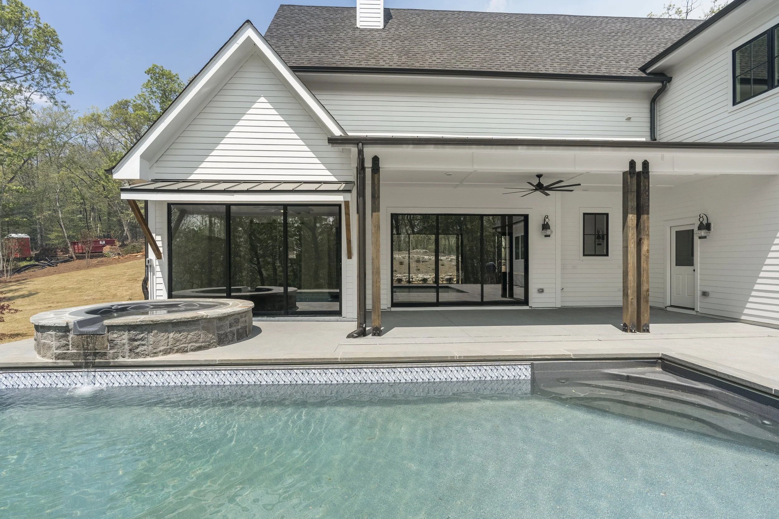 Backyard with a swimming pool, hot tub, and a modern house with a large covered patio area. The house has white siding, large glass doors, and a ceiling fan. There are trees and a blue sky in the background.