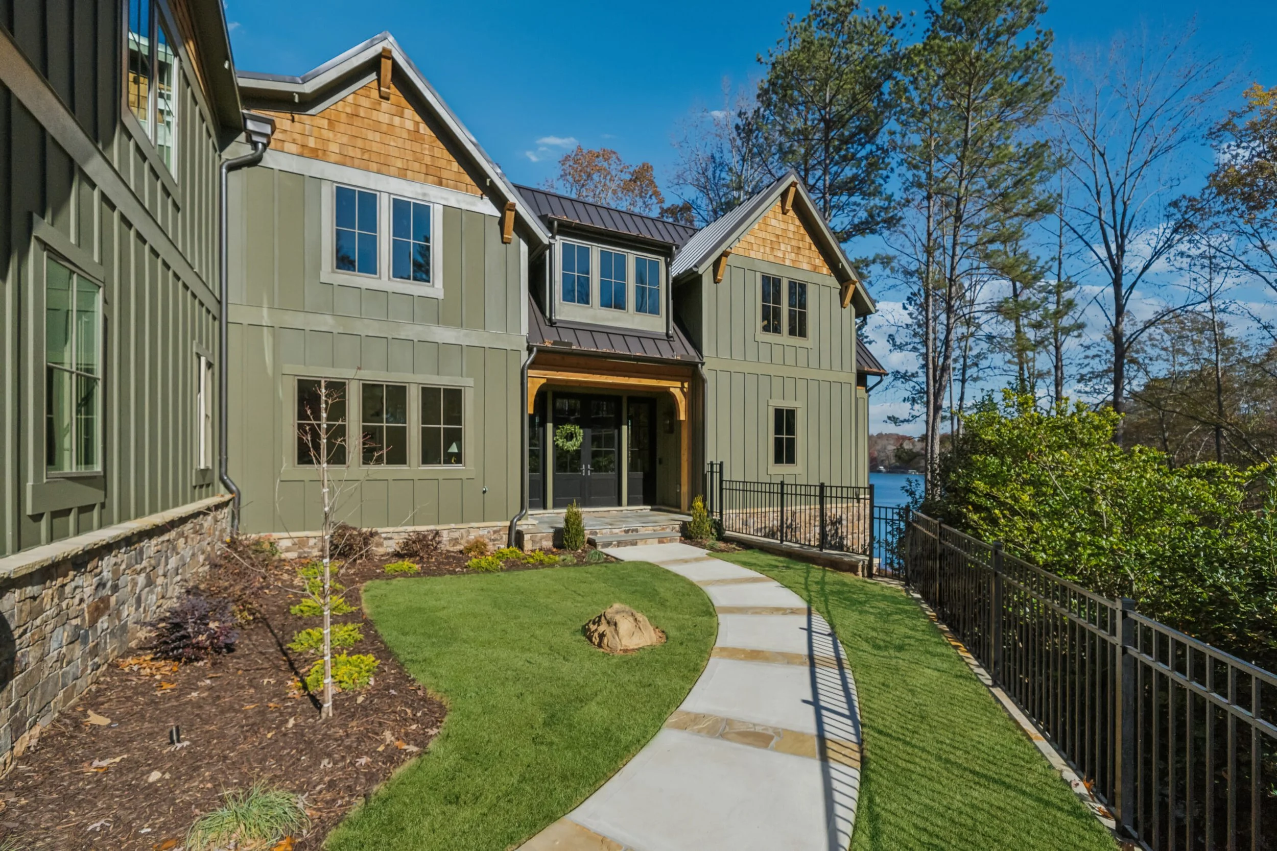 A newly constructed house with light green exterior siding, large windows, and a stone foundation. There is a small front yard with a curved concrete pathway, a young tree, and some landscaped bushes. The house has a dark metal roof and is surrounded