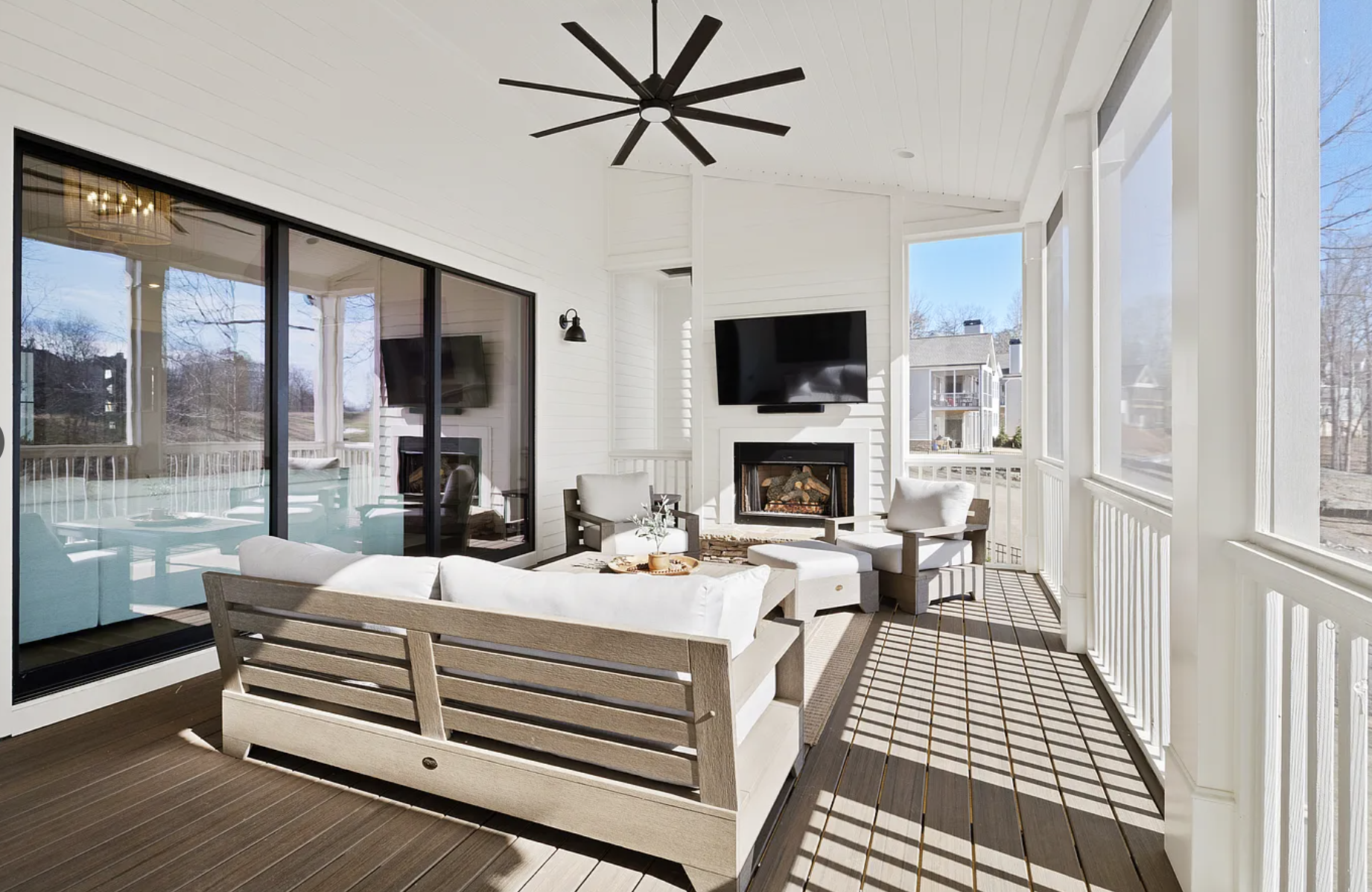Bright porch with white walls, black ceiling fan, and outdoor furniture including cushioned chairs and a coffee table. Large windows show a neighboring house and trees, with sunlight casting shadows on the floor.