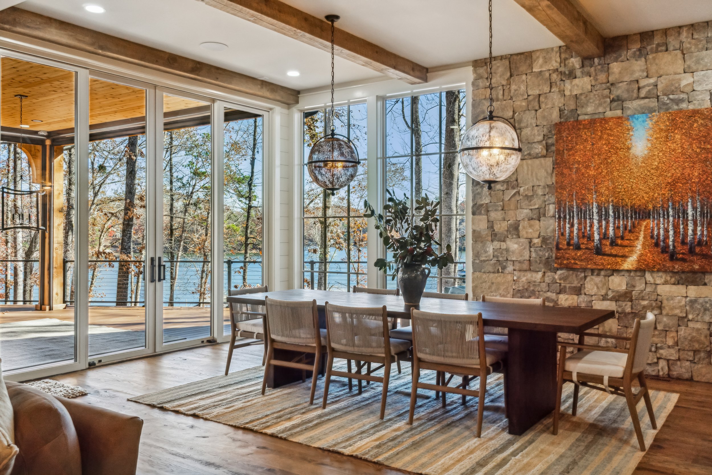 A dining area with a wooden table and chairs, large windows showing trees outside, stone accent wall with a colorful abstract painting, two hanging metal pendant lights, with a view of a covered porch and forest beyond.