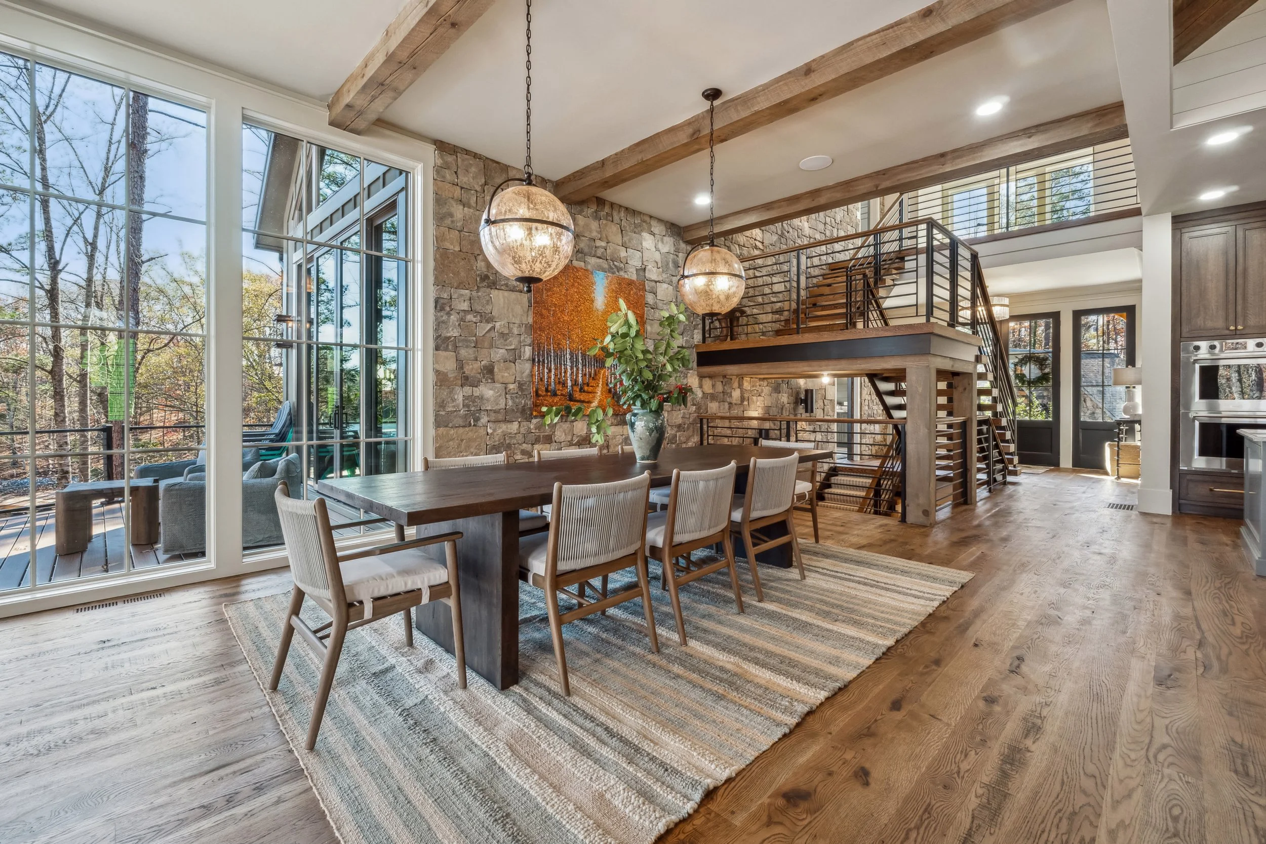 Modern dining room with large windows, wooden beams, stone accent wall, circular pendant lights, a dining table with chairs, potted plant, staircase, and hardwood floors.