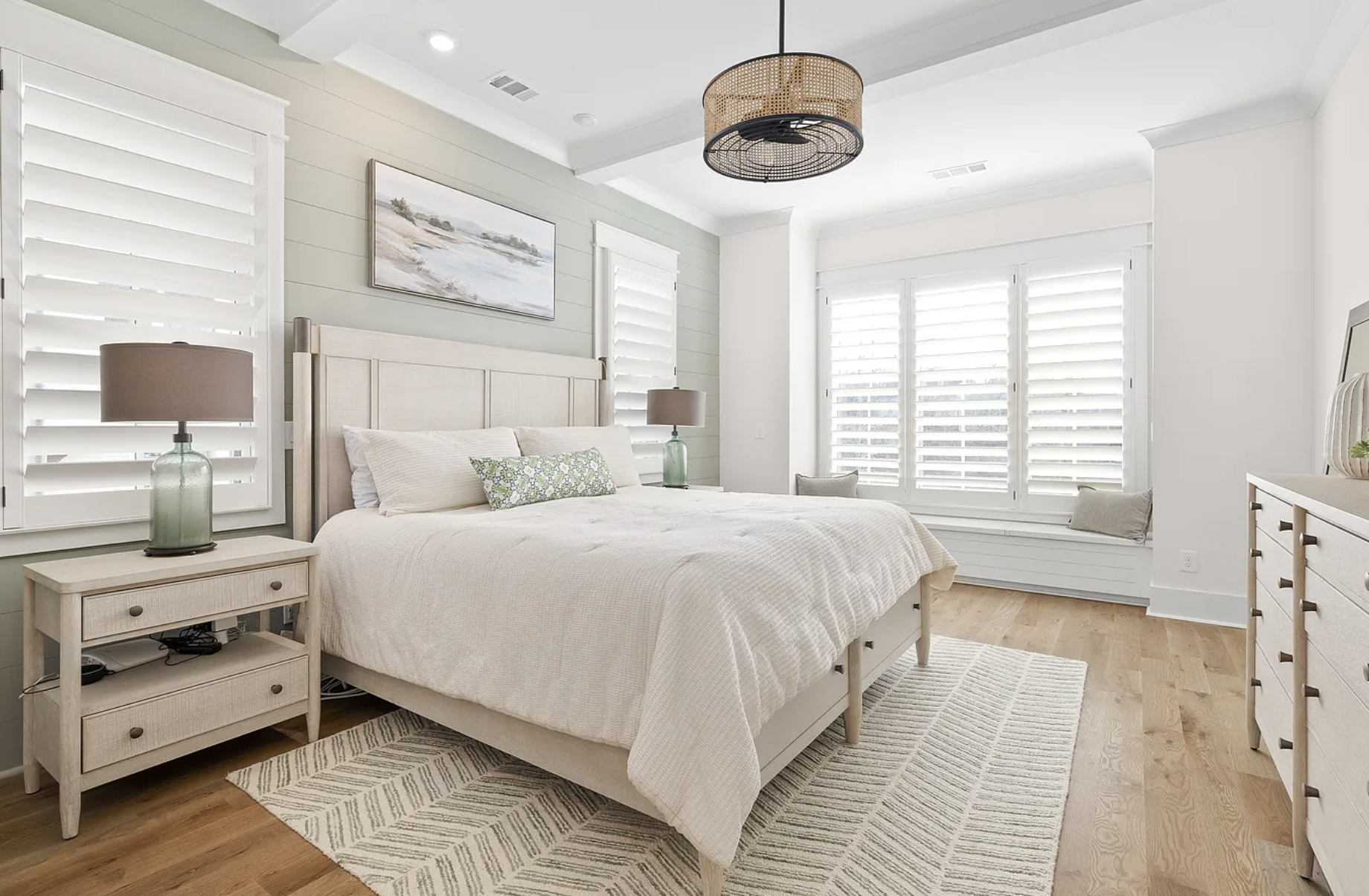 A bright, white bedroom with a large bed, nightstands, lamps, and windows with white shutters.