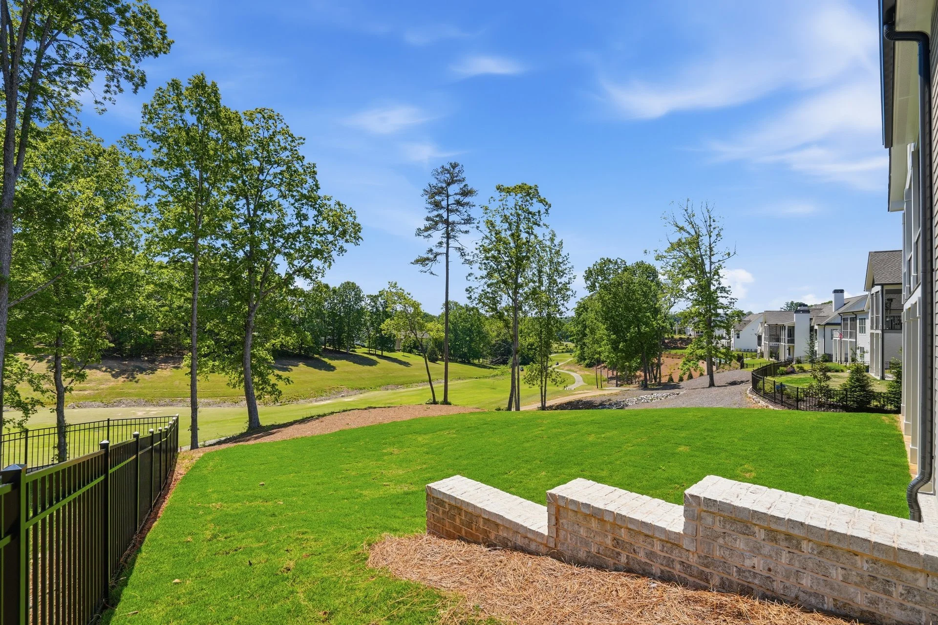 Backyard with green lawn, brick patio, black metal fence, trees, a small pond, and a walking path, with white residential buildings in the background under a blue sky.