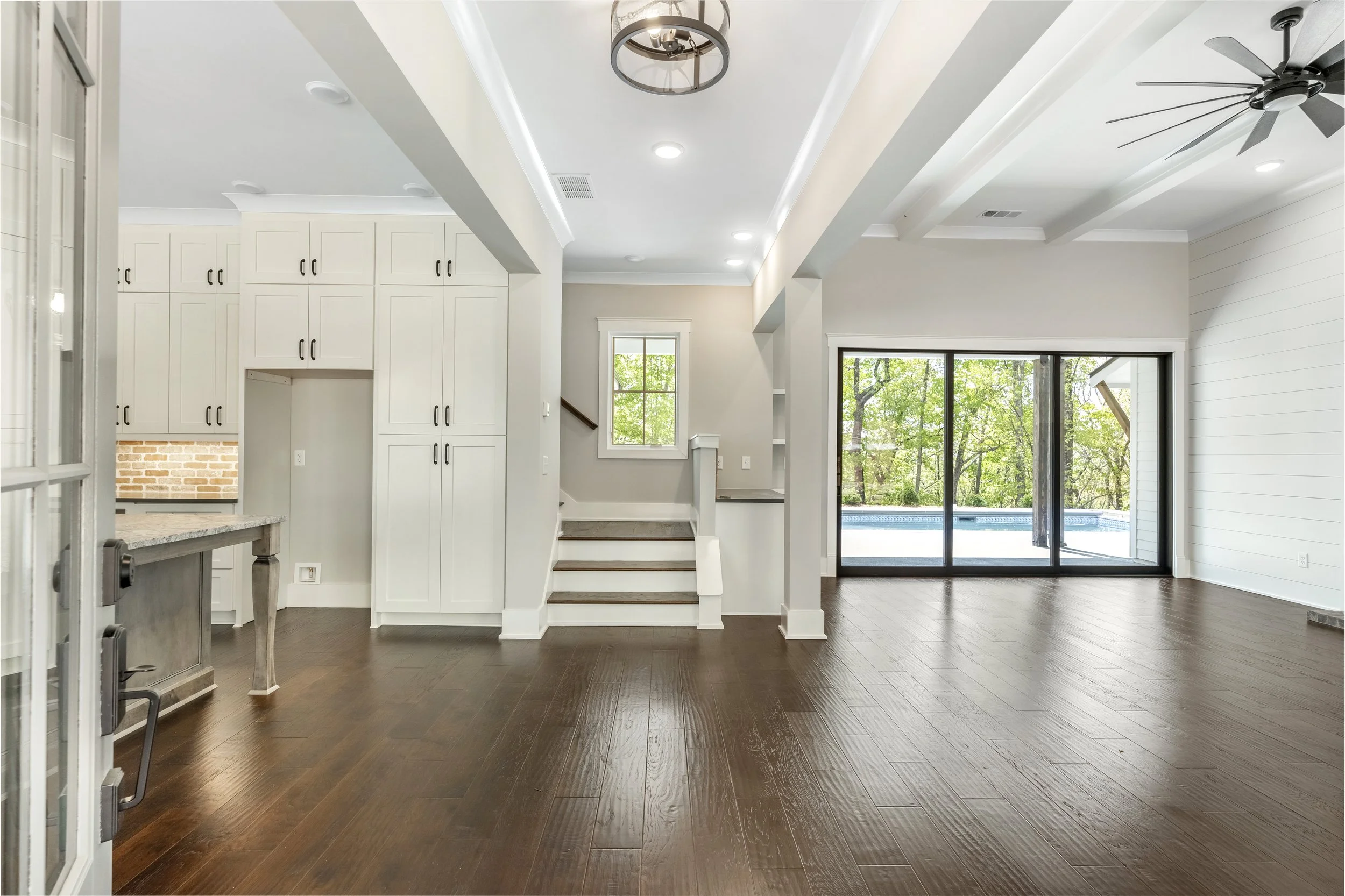 Interior view of a modern home living area with hardwood floors, white walls, a staircase, large sliding glass doors leading to a pool, and ceiling fans.