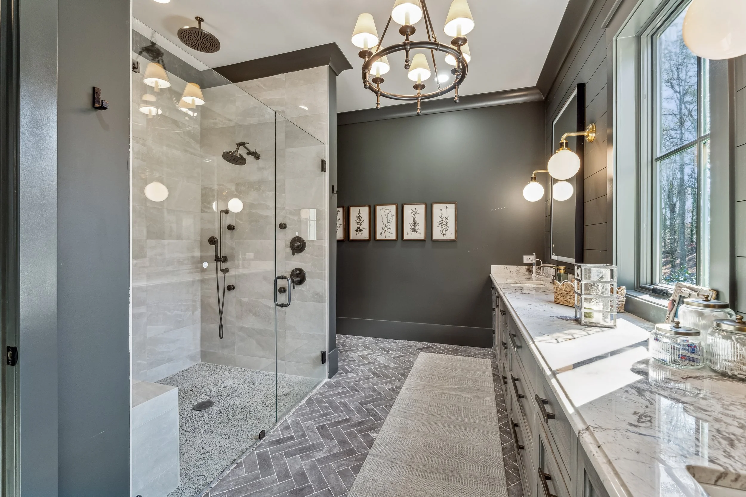 Modern bathroom with a glass-enclosed shower featuring multiple shower heads and dark fixtures. The room has a dark green wall, art prints of botanical illustrations, a marble countertop with bathroom essentials, and a window letting in natural light