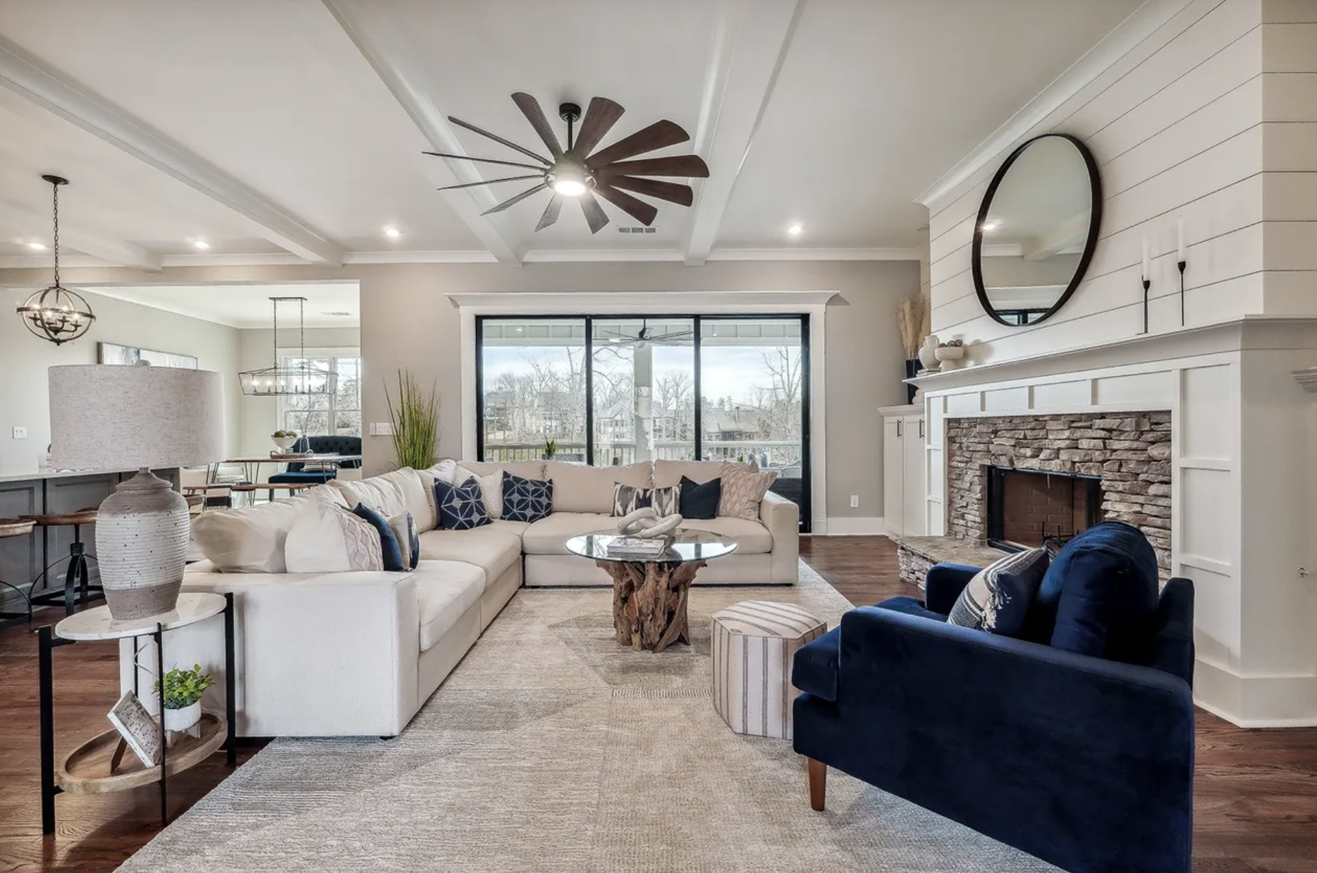 A living room with a white sectional sofa decorated with navy and patterned throw pillows, a round glass coffee table with a wood base, a blue armchair, a fireplace with a white mantel and shiplap wall above, a large round mirror, and a sliding glass