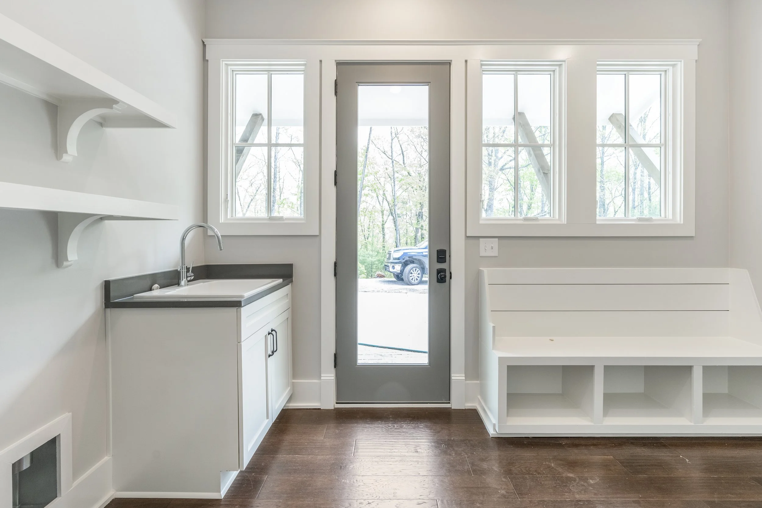 Interior of a minimalistic kitchen with white cabinets, a sink, and large windows and door overlooking trees and a parked car outside.