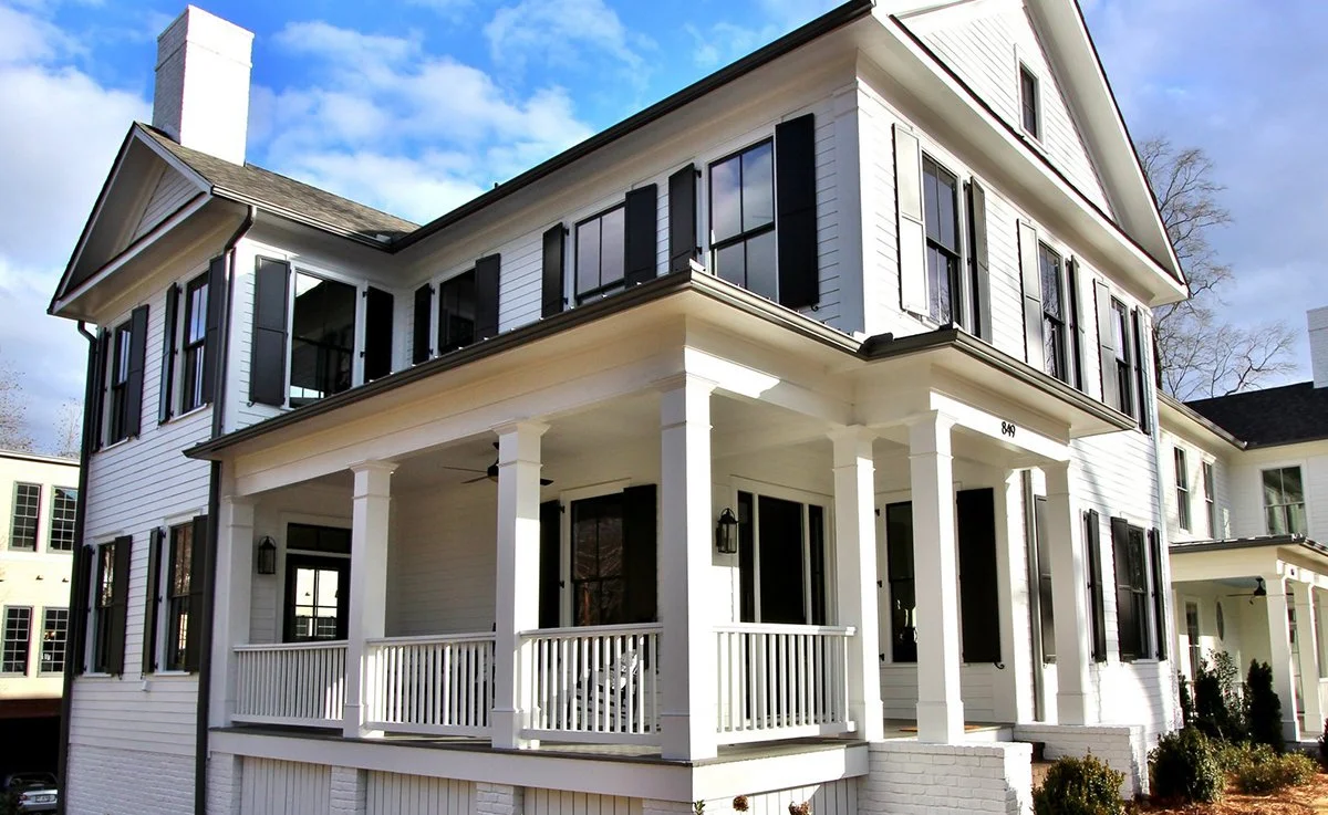 A large white multi-story house with black shutters, a covered porch, and a gabled roof under a partly cloudy sky.