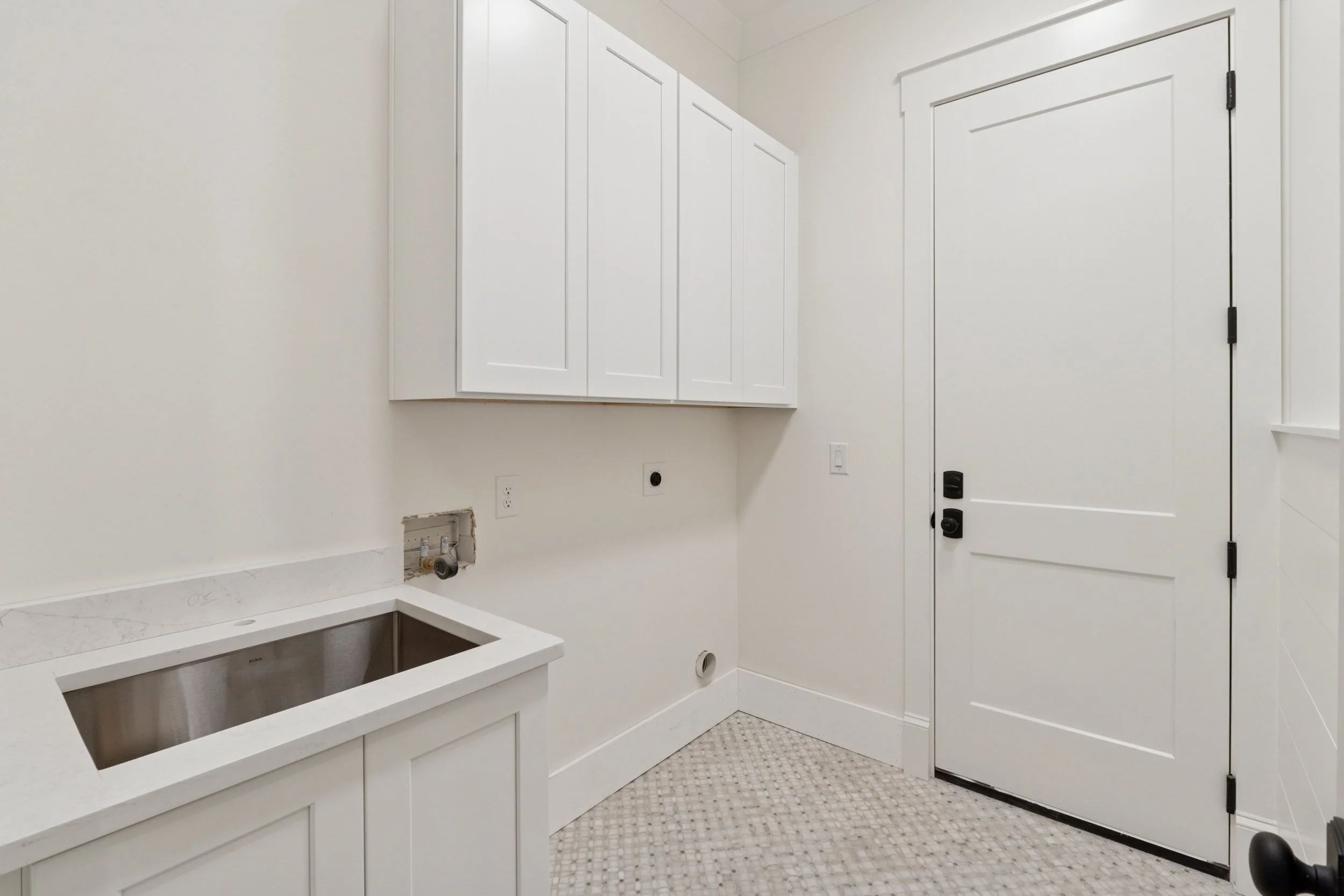 Empty laundry room with white cabinets, a sink, and a door, with plumbing and electrical outlets visible.