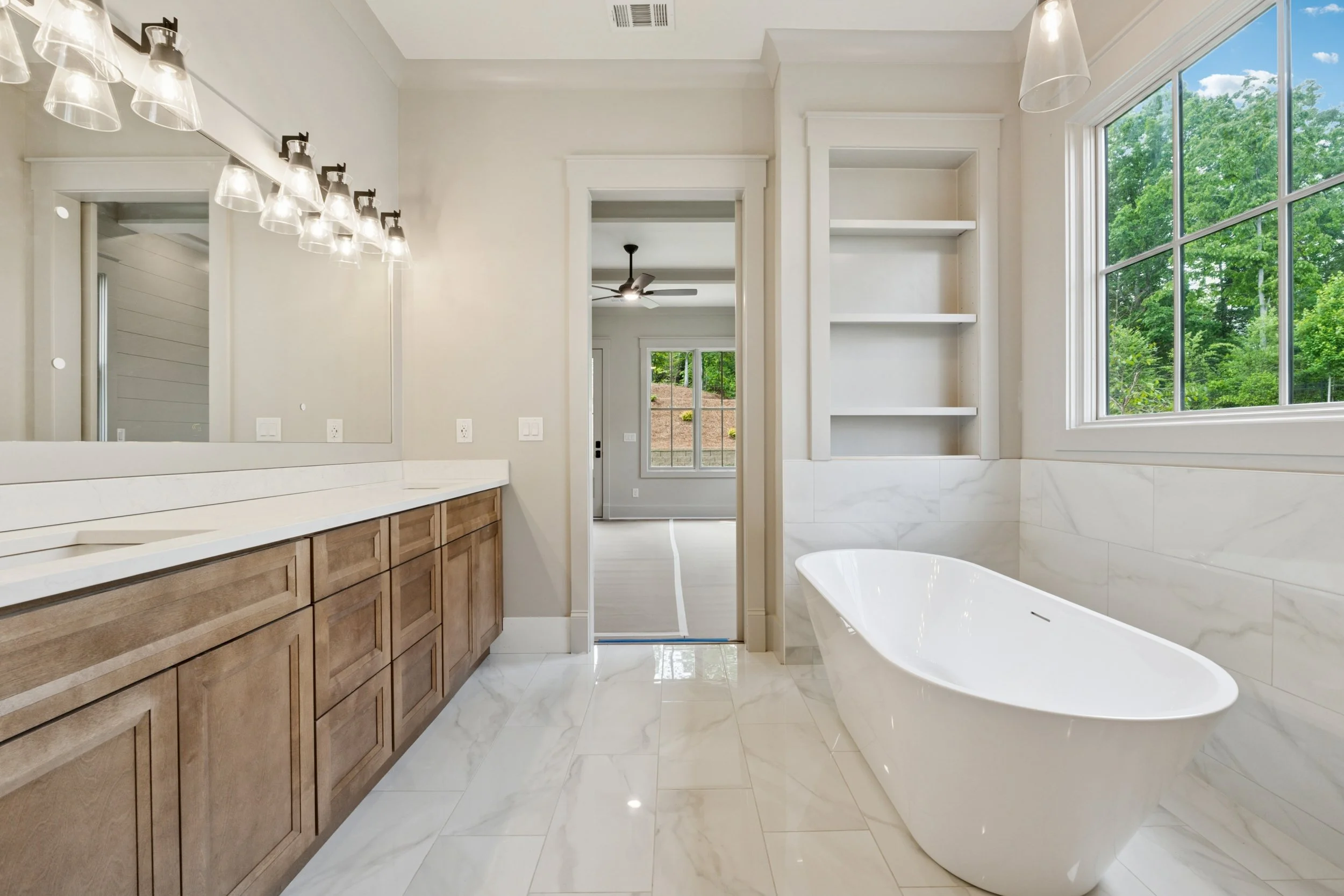 Modern bathroom with a large bathtub, wooden vanity with multiple drawers, marble flooring, built-in white shelves, large window with a view of greenery, ceiling fan, and stylish lighting fixtures.