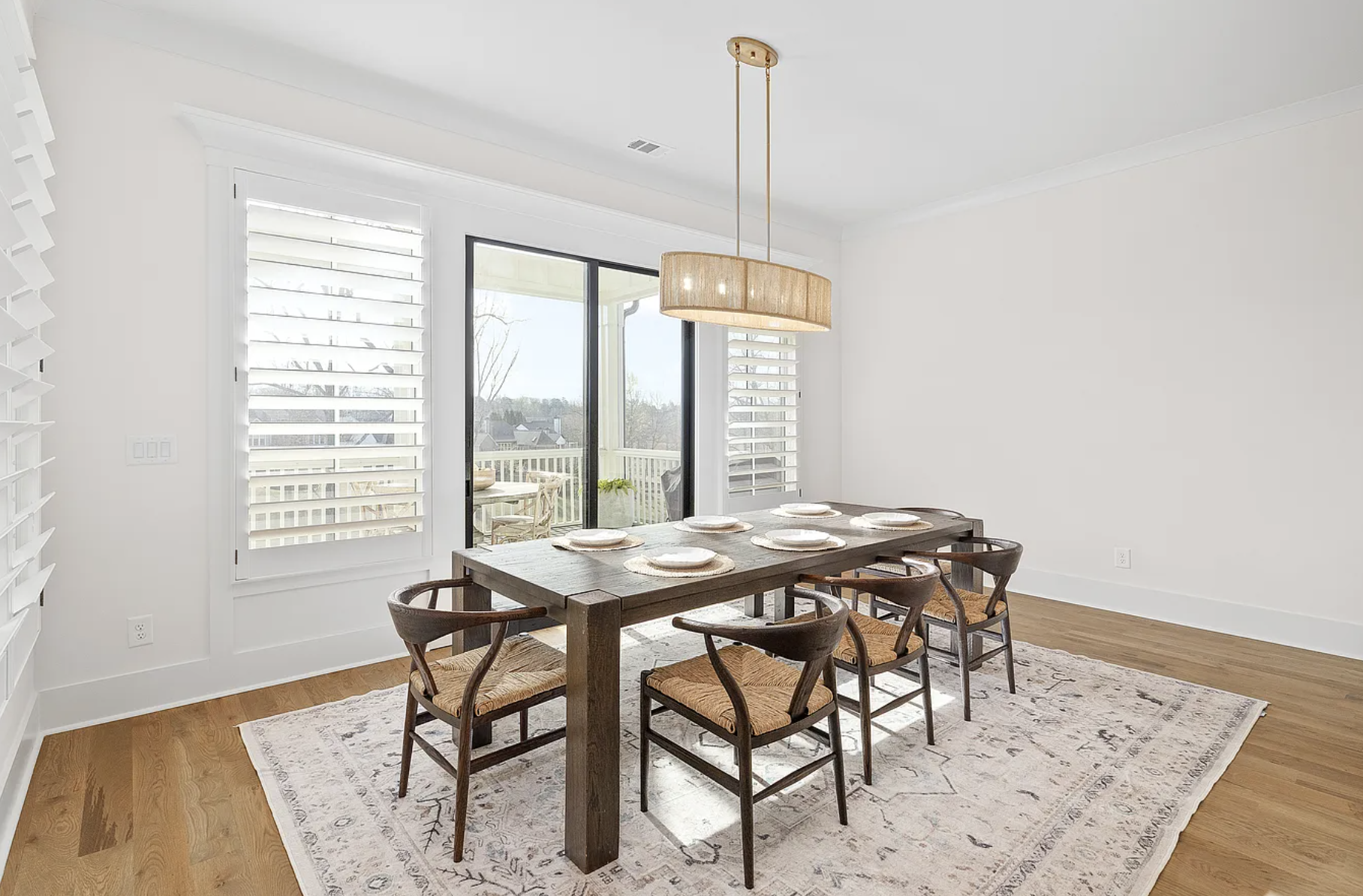A dining room with a wooden table, six chairs with woven seats, a light fixture hanging above, large sliding glass door opening to a balcony, and windows with plantation shutters, all with white walls and hardwood floors.