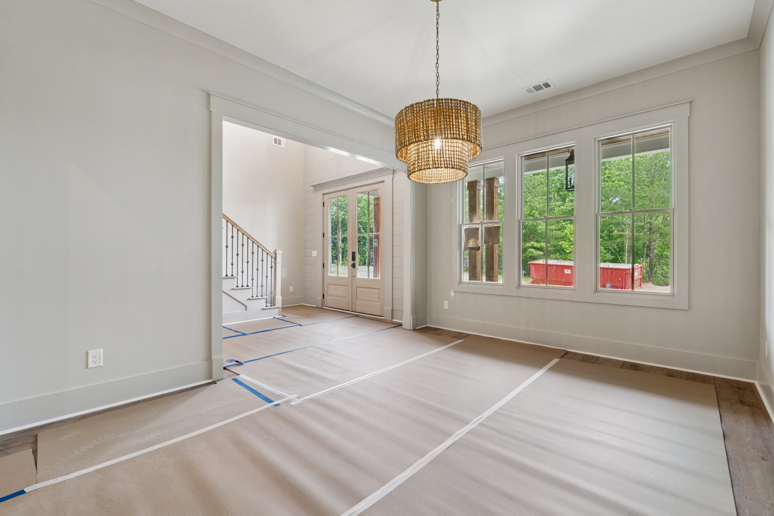 Interior view of a house under renovation, with tan paper on the floor, blue painter's tape outlining areas, large windows showing green trees outside, a chandelier hanging from the ceiling, and an entry door at the front.