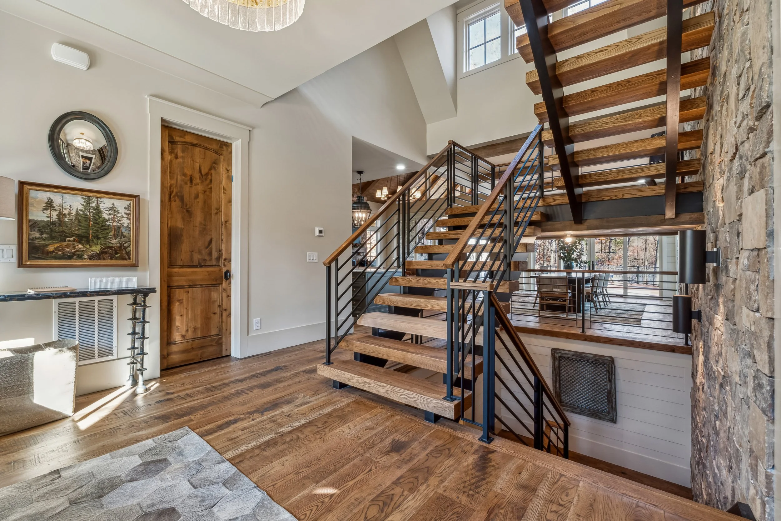 Interior view of a modern home with wooden and stone accents, staircase with wooden treads and black metal railing, chandelier, framed artwork on the wall, and large windows showing outdoor scenery.