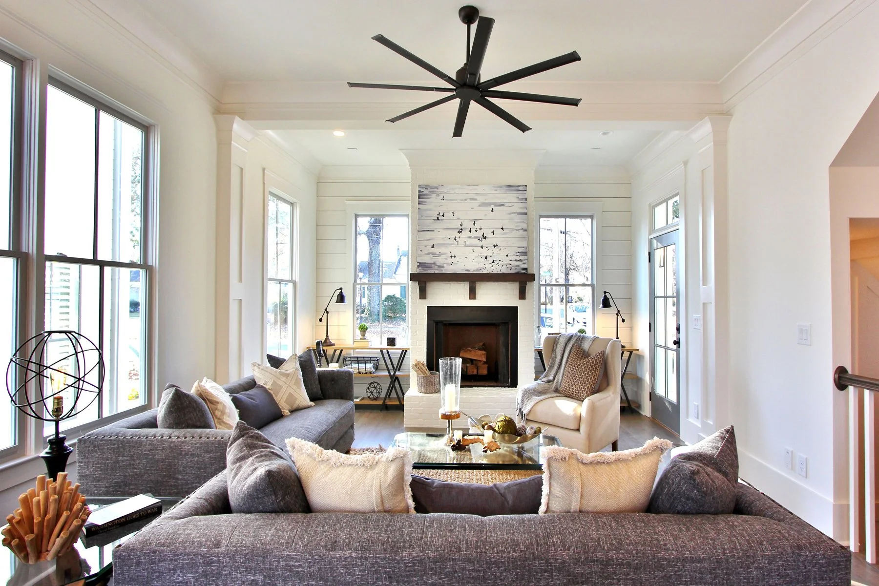 Bright living room with large windows, a fireplace with a white and gray wooden mantle, and modern furniture including a gray sofa, beige armchair, and black side tables with lamps. The room features a black ceiling fan and decorative items on coffee