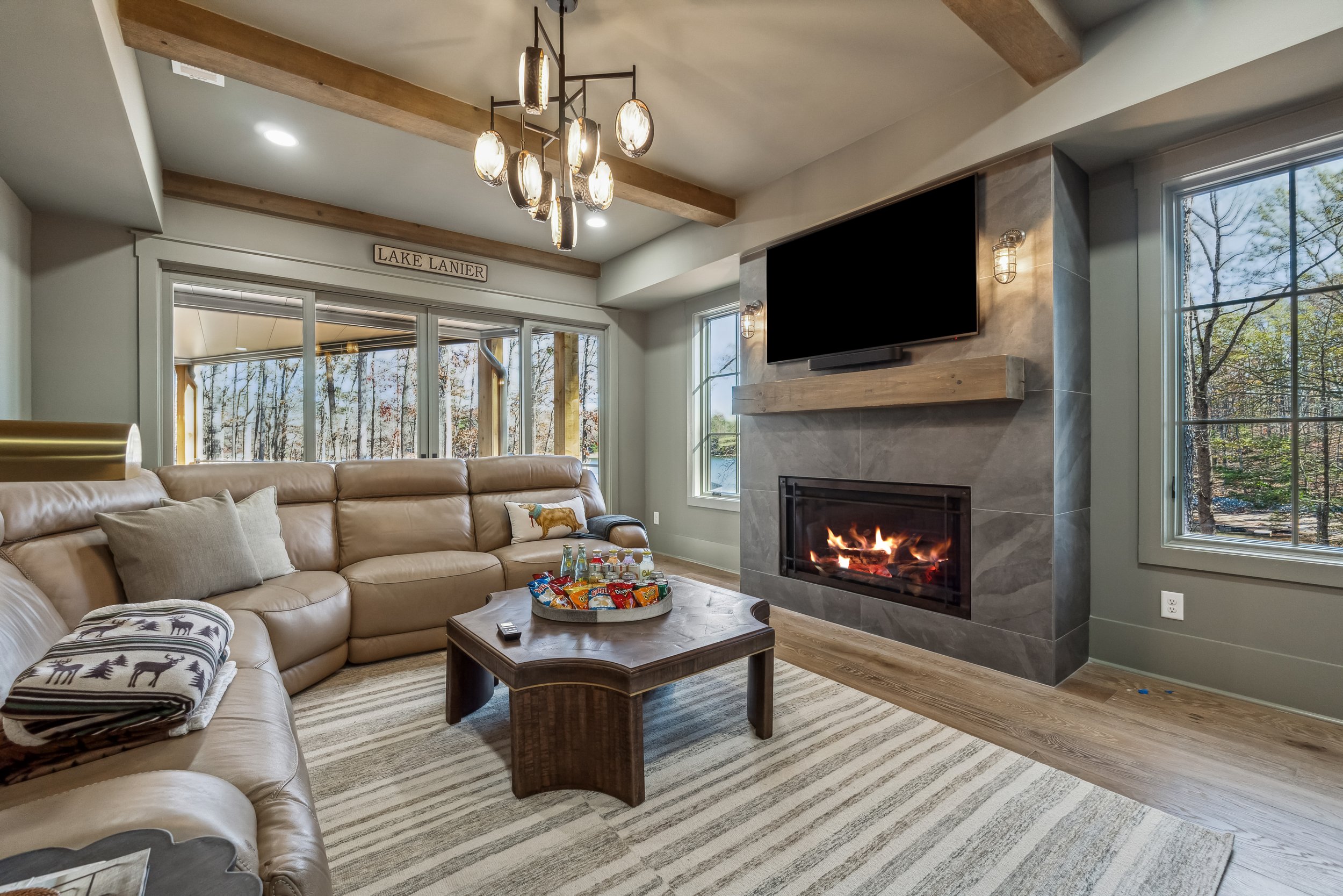 Living room with beige sectional sofa, wooden coffee table with snacks, fireplace, TV above fireplace, large windows showing trees outside, and a modern chandelier with hanging lights.