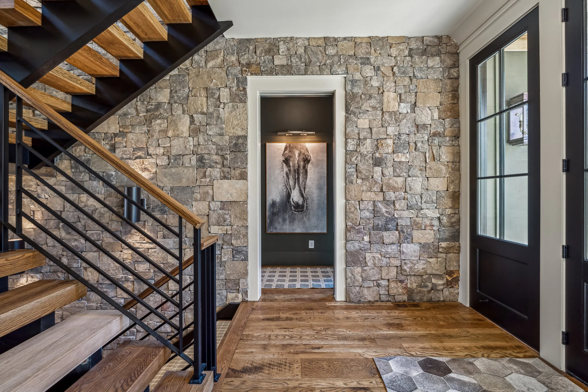 Interior view of a modern home entry with a stone accent wall, staircase with wooden steps and black metal railing, hardwood floors, a framed artwork, and front door with glass panes.