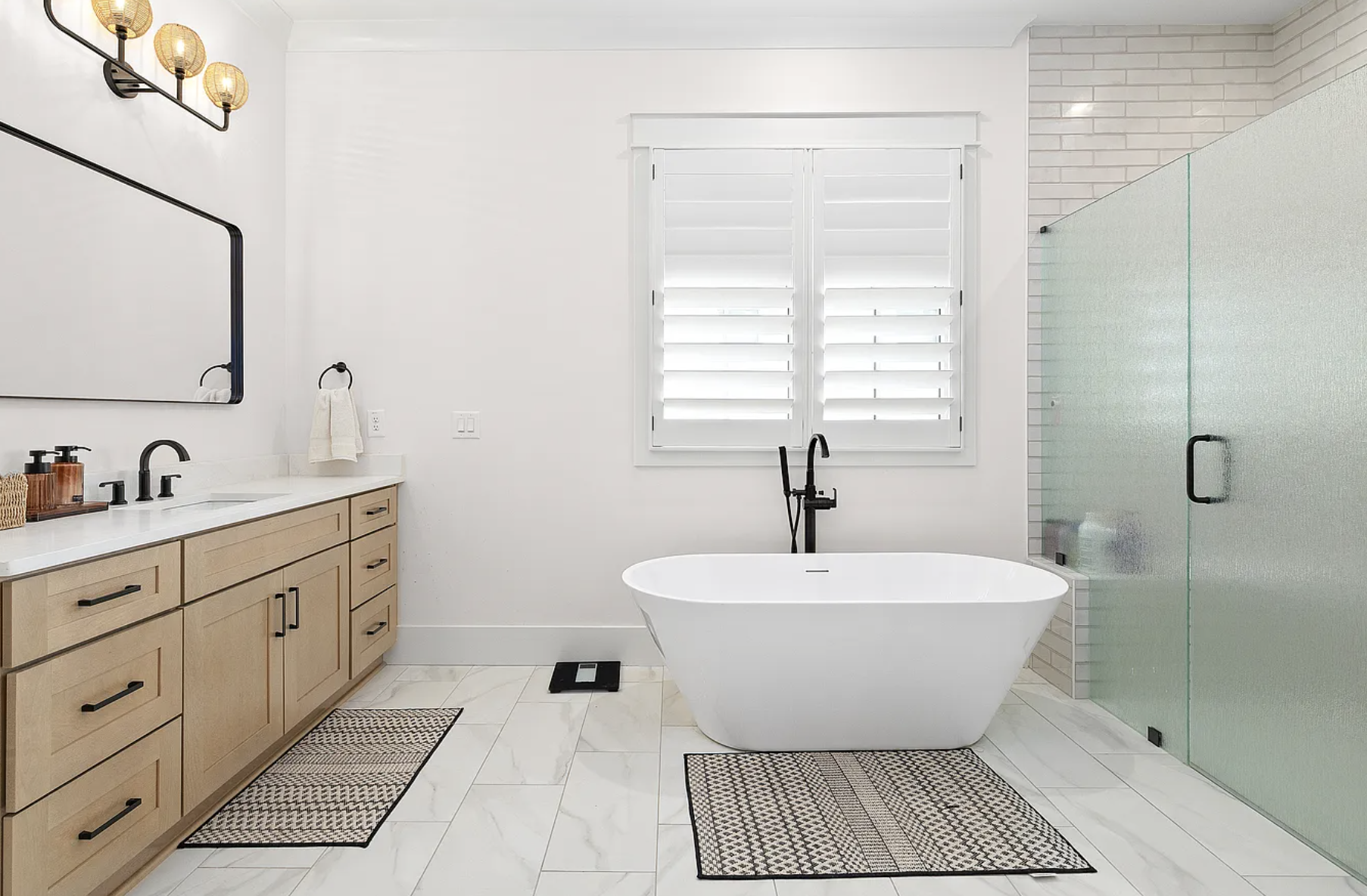 Modern bathroom with white walls, a bathtub beneath a window with white shutters, light wood vanity with black hardware, black fixtures, glass shower enclosure, and beige marble flooring with patterned rugs.