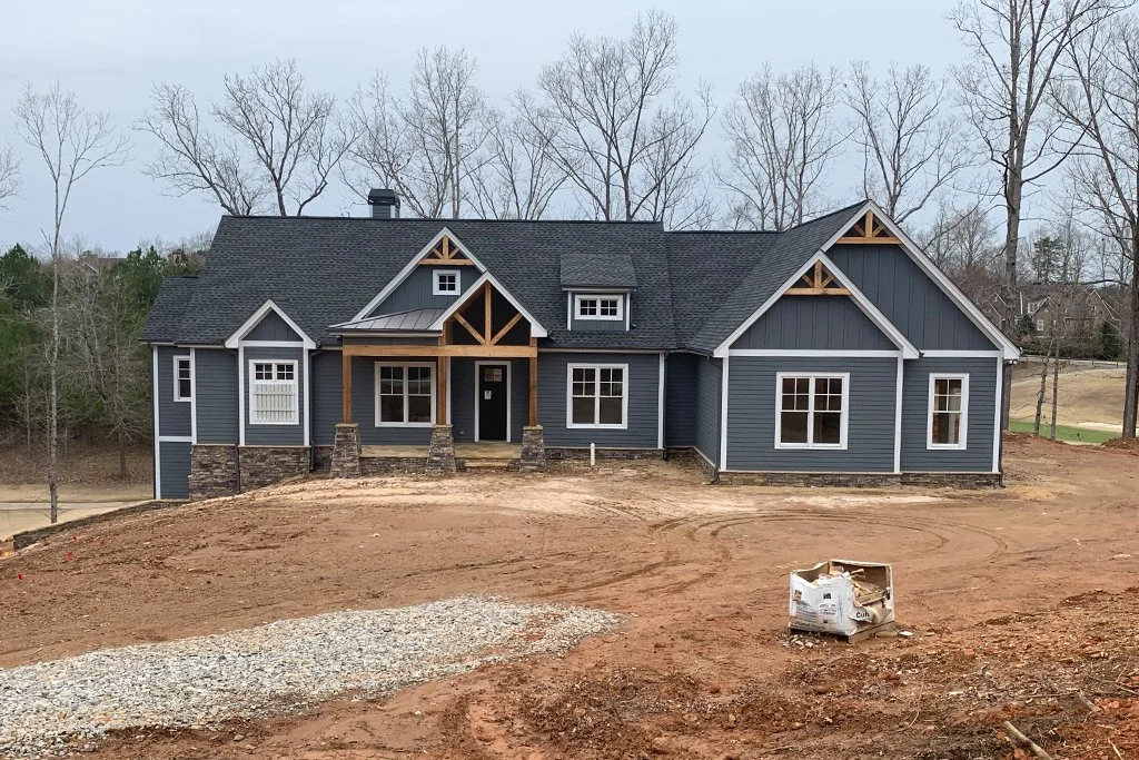 A newly constructed house with a dark blue exterior, white-trimmed windows, and a black roof, situated on a dirt plot of land with some construction materials in the foreground, and leafless trees in the background.